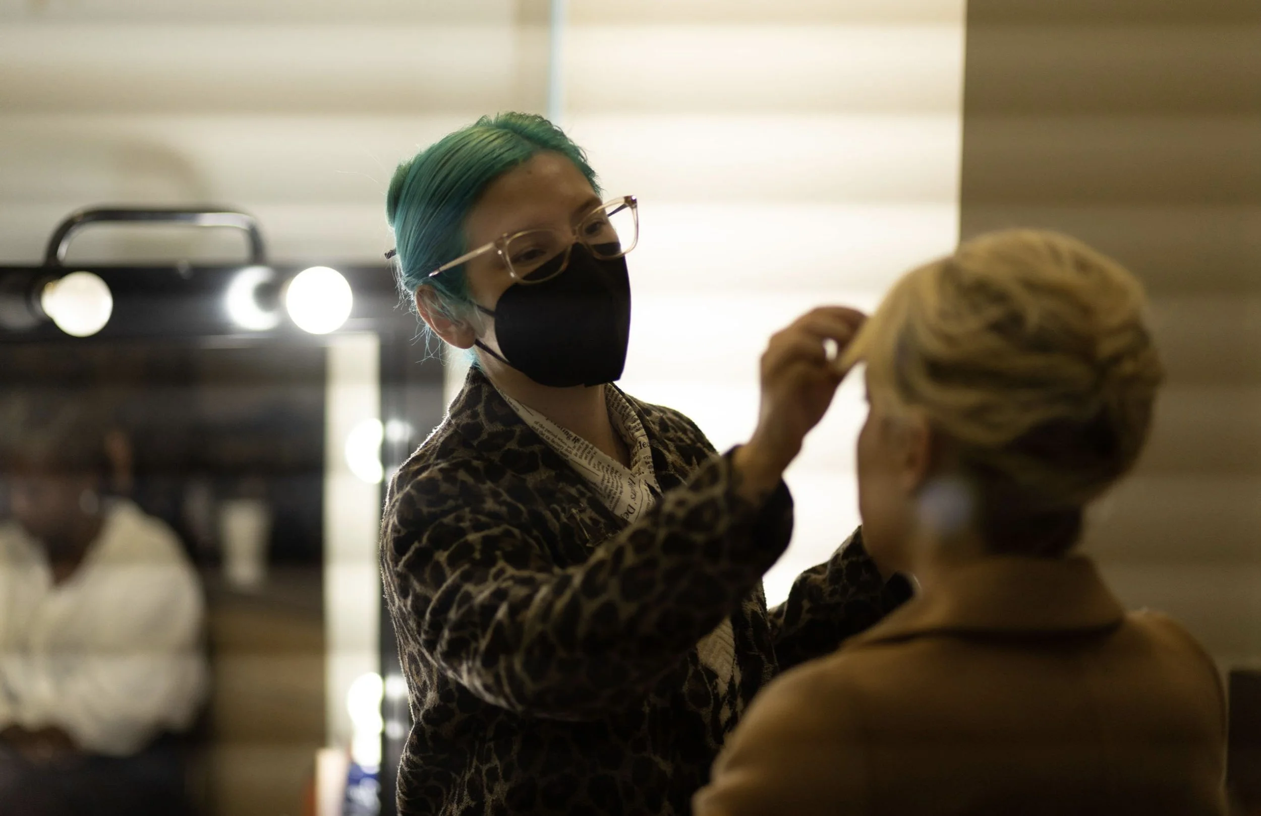 A makeup artist is applying makeup to a woman's face in a salon or beauty studio. The makeup artist has teal-green hair, glasses, and a black face mask, wearing a leopard-print jacket. The woman has blonde hair styled in an updo and is wearing a beig