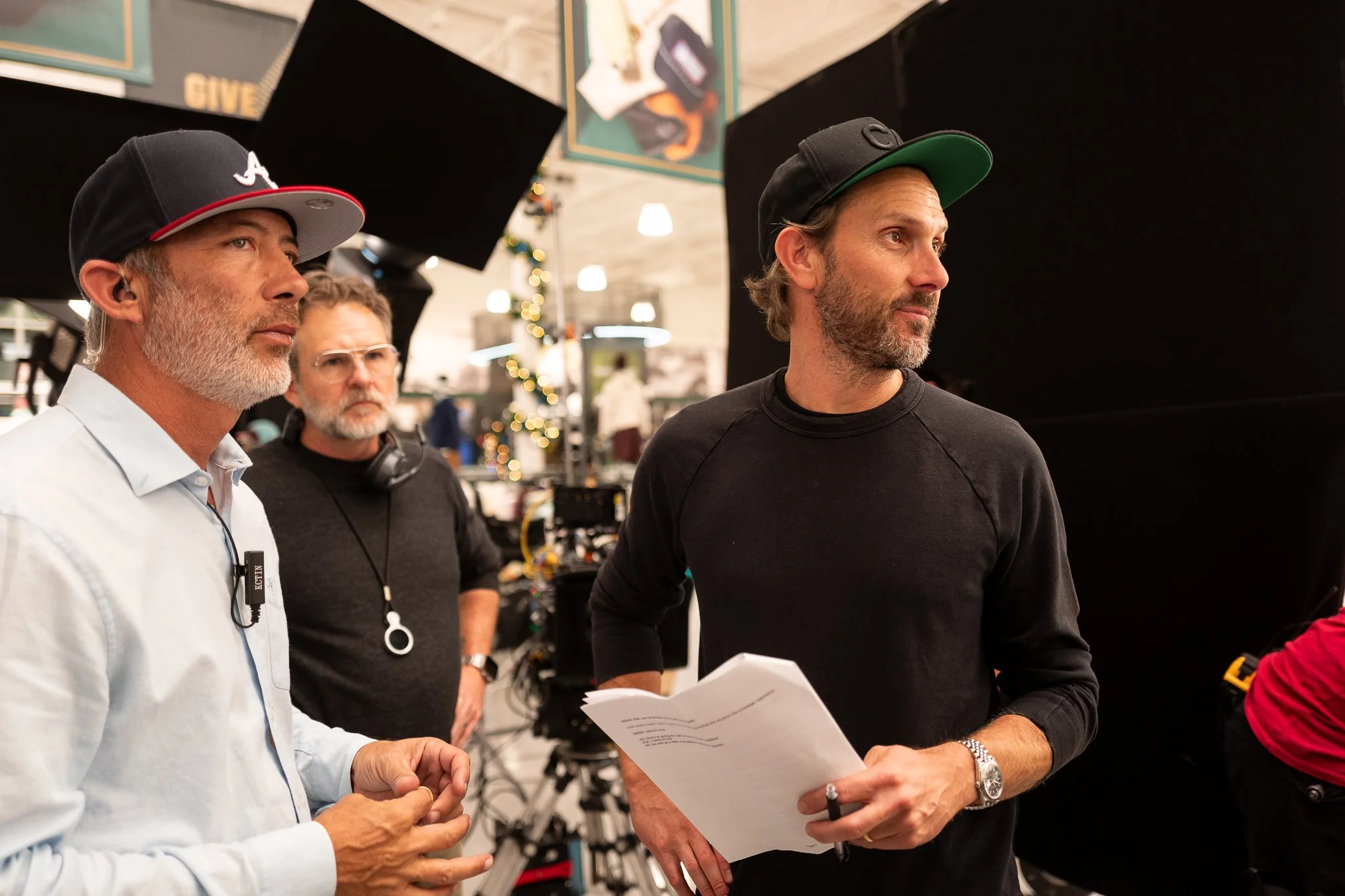 Three men on a film set, one wearing a black cap and black shirt holding papers, the second with glasses and headset, and the third with a university cap, standing near a camera and equipment.
