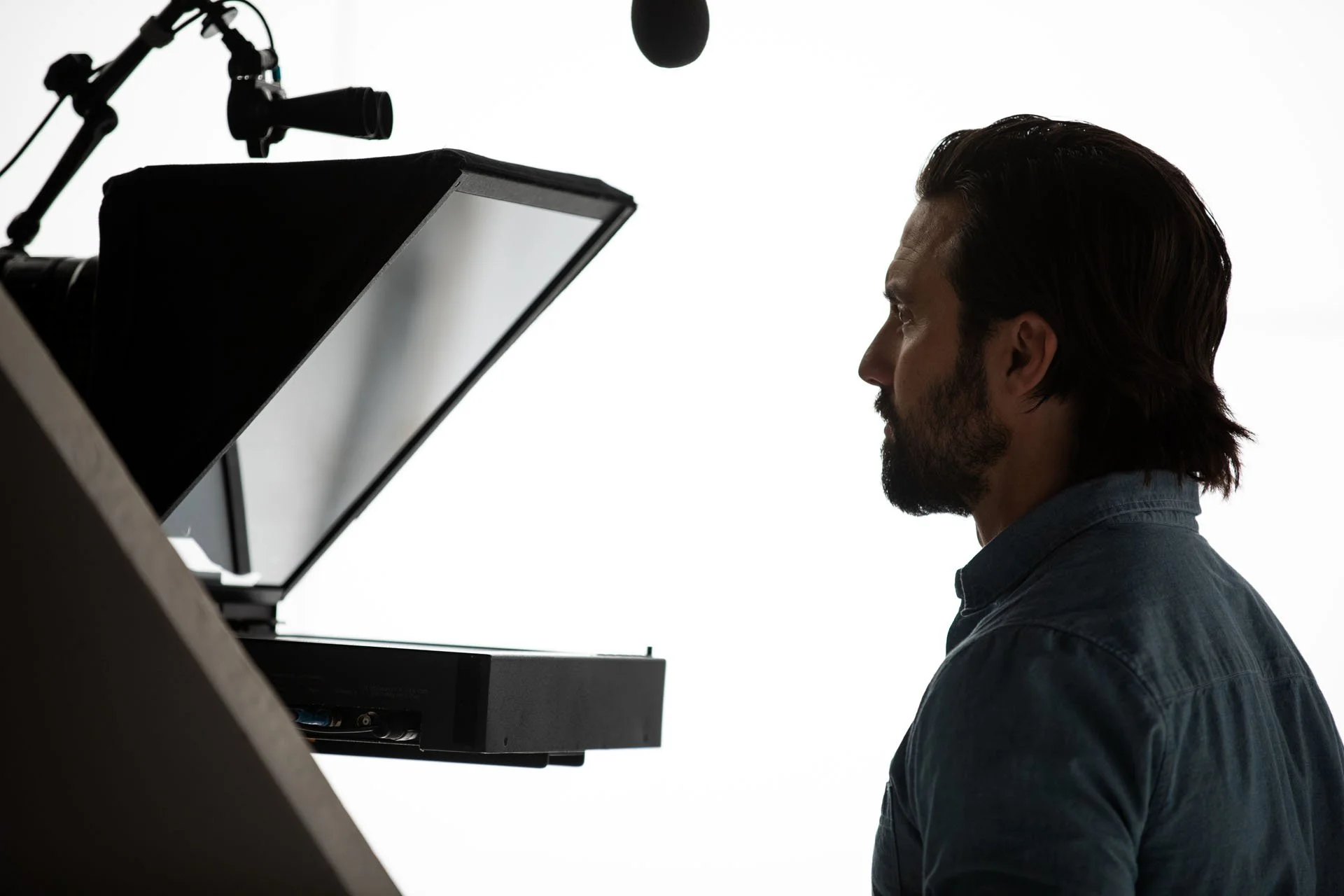 A silhouette of a man with dark hair and a beard, sitting in front of a laptop with a microphone hanging above, against a bright white background.