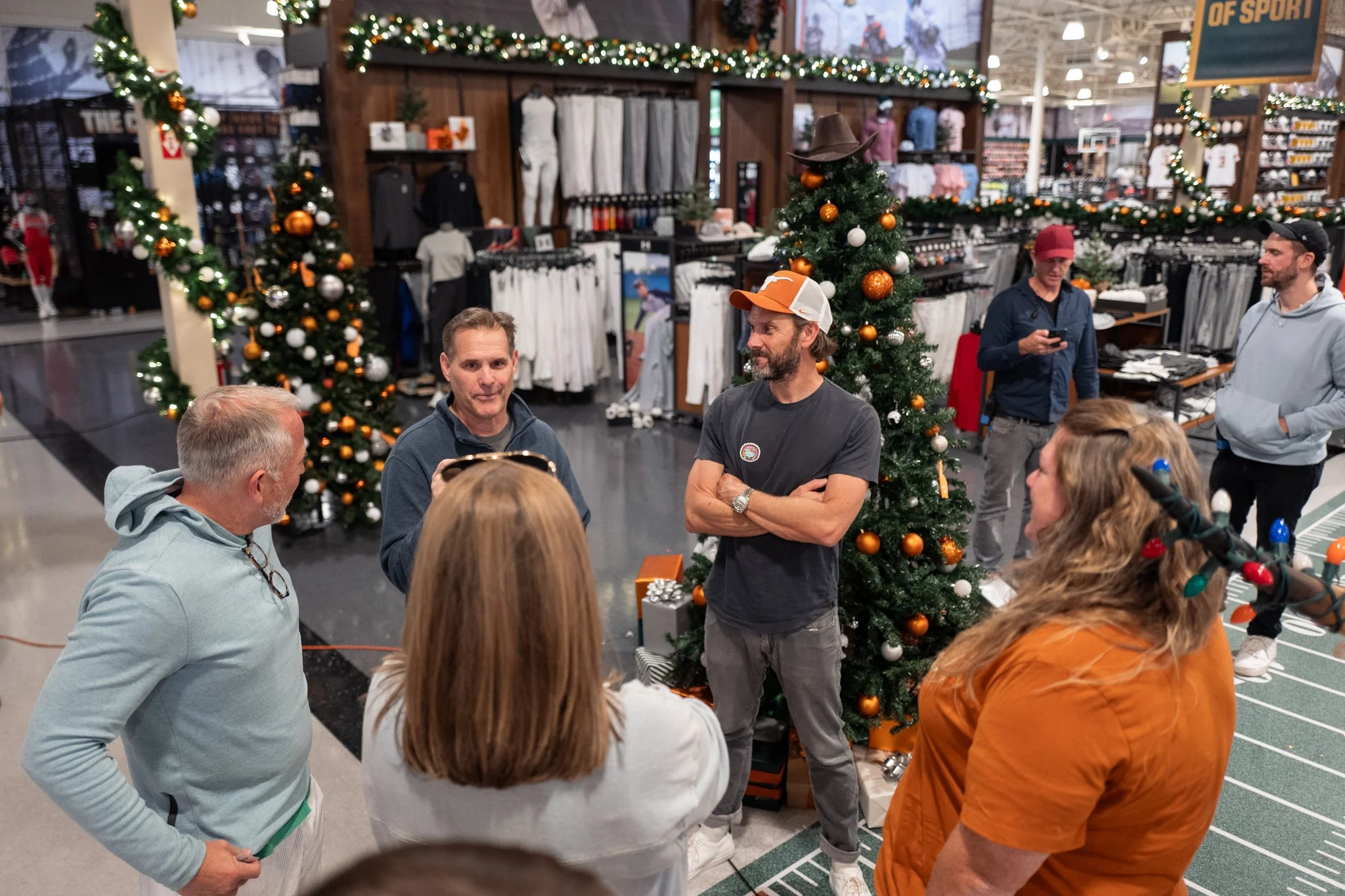 Group of people gathered around a man speaking at a holiday event in a store decorated with Christmas trees and garlands.