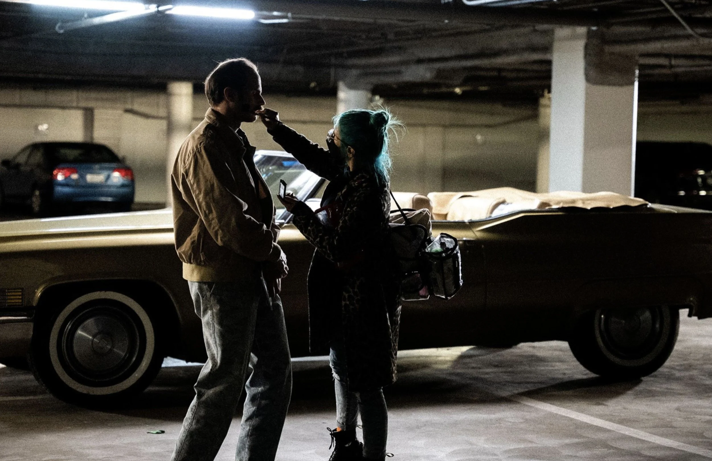 A woman with blue hair is applying makeup or touching up lipstick to a man in a parking garage, with a vintage car and several other parked cars in the background.