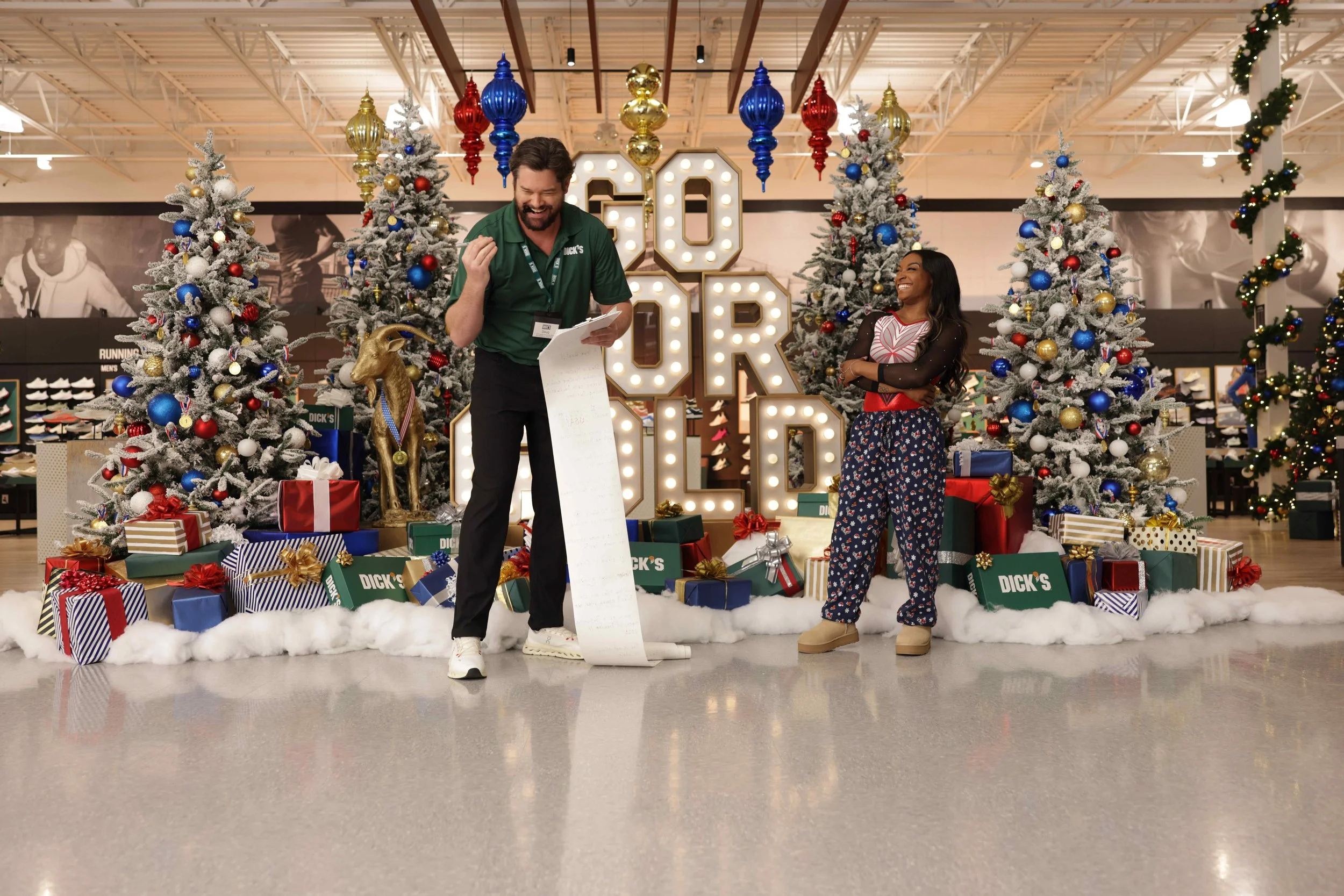 Two people standing in front of Christmas trees and holiday decorations in a store, with one person smiling and the other looking amused, amid presents and holiday decor.
