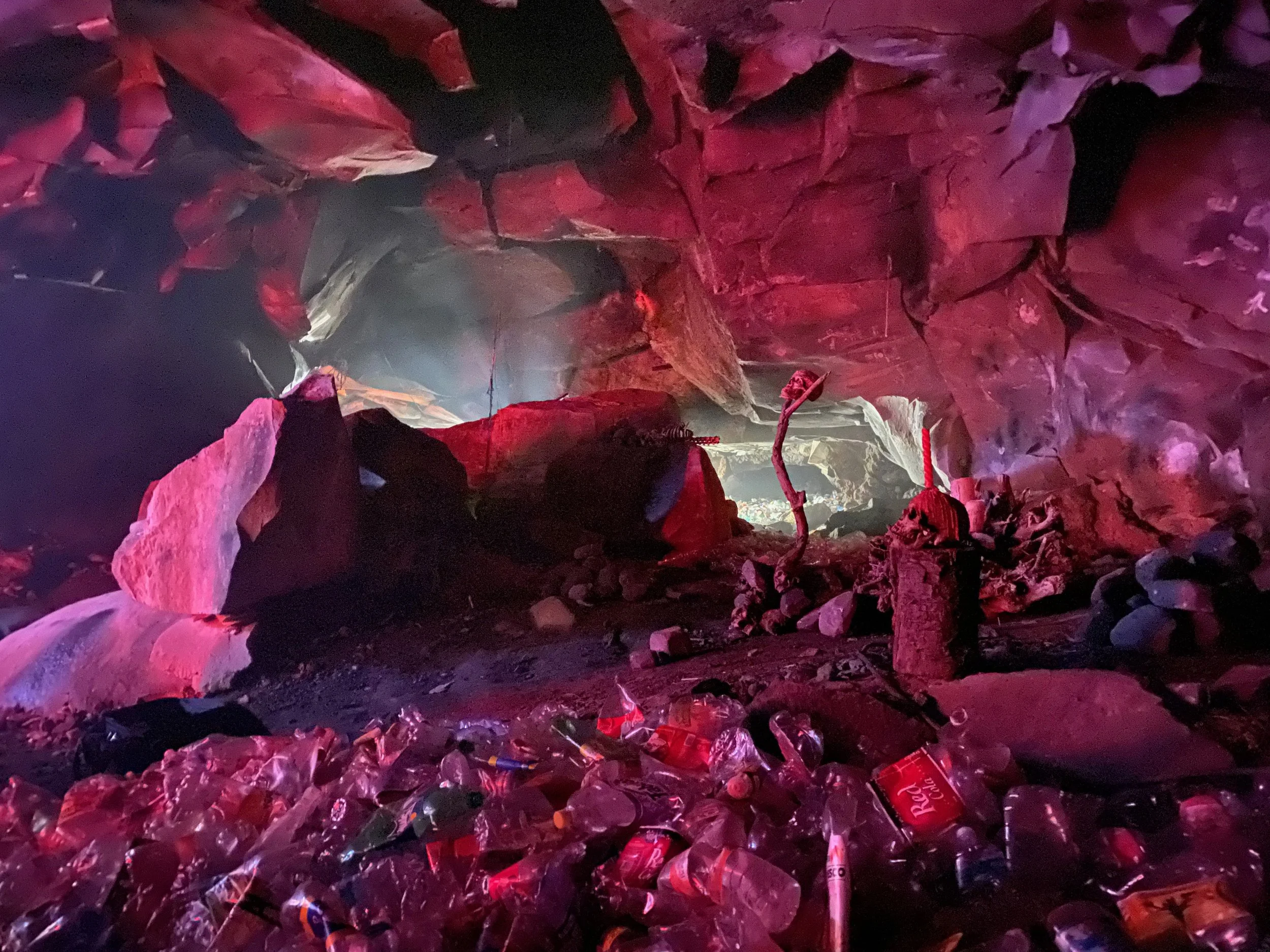 Inside a cave with rocks and stalagmites, illuminated by red and purple lighting, with a large amount of empty plastic bottles and cans scattered on the ground.