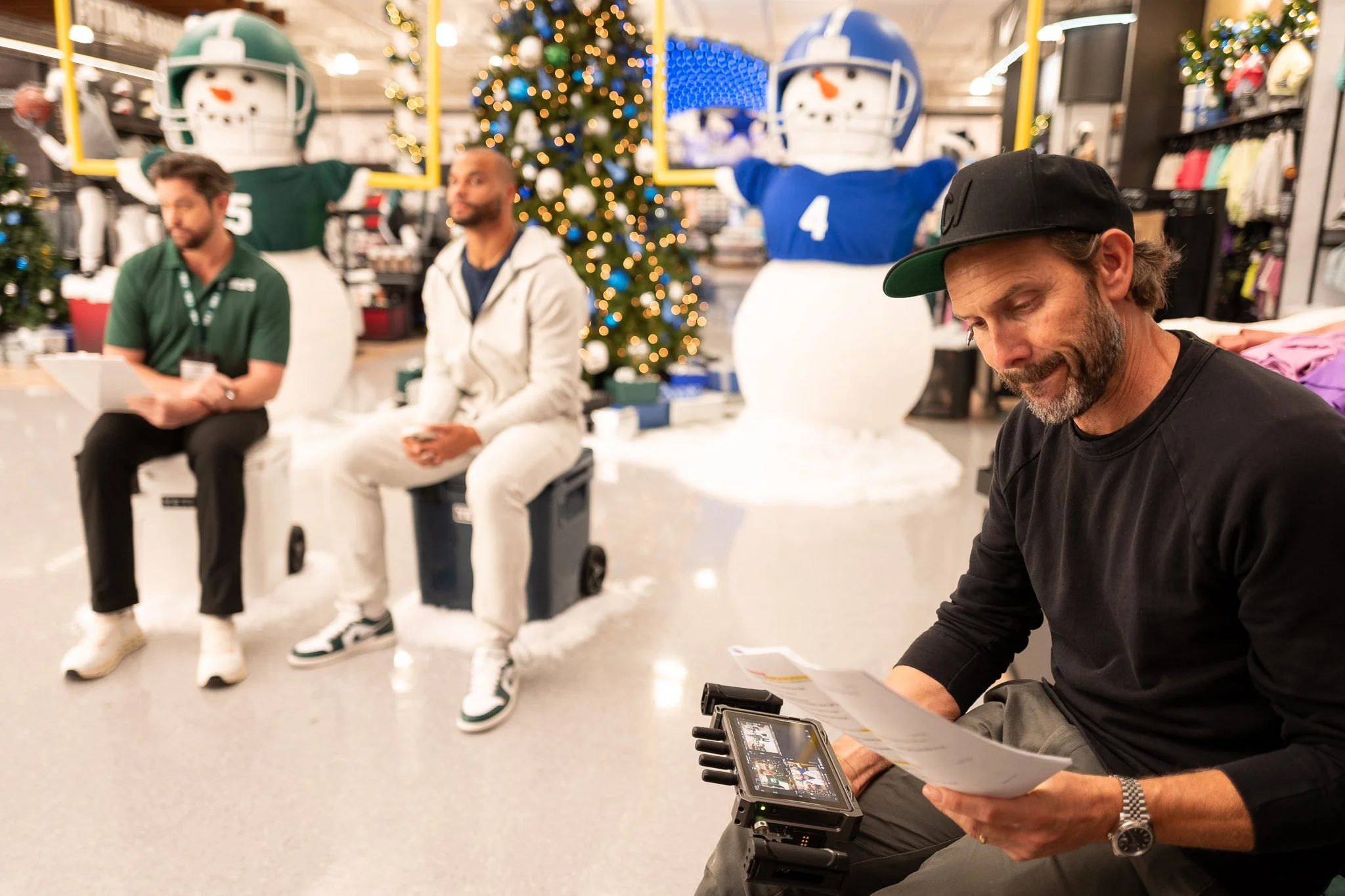 People sitting in a store decorated with Christmas trees and snowmen wearing football helmets, during a holiday event or interview.