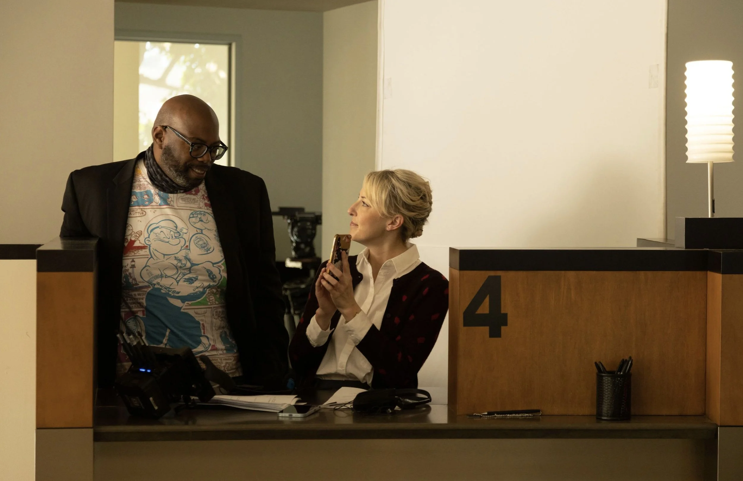 A man and woman inside a reception area, engaged in conversation. The woman holds a phone and is smiling at the man. They are near a numbered desk labeled '4' with a cup of pens and some documents on the counter.