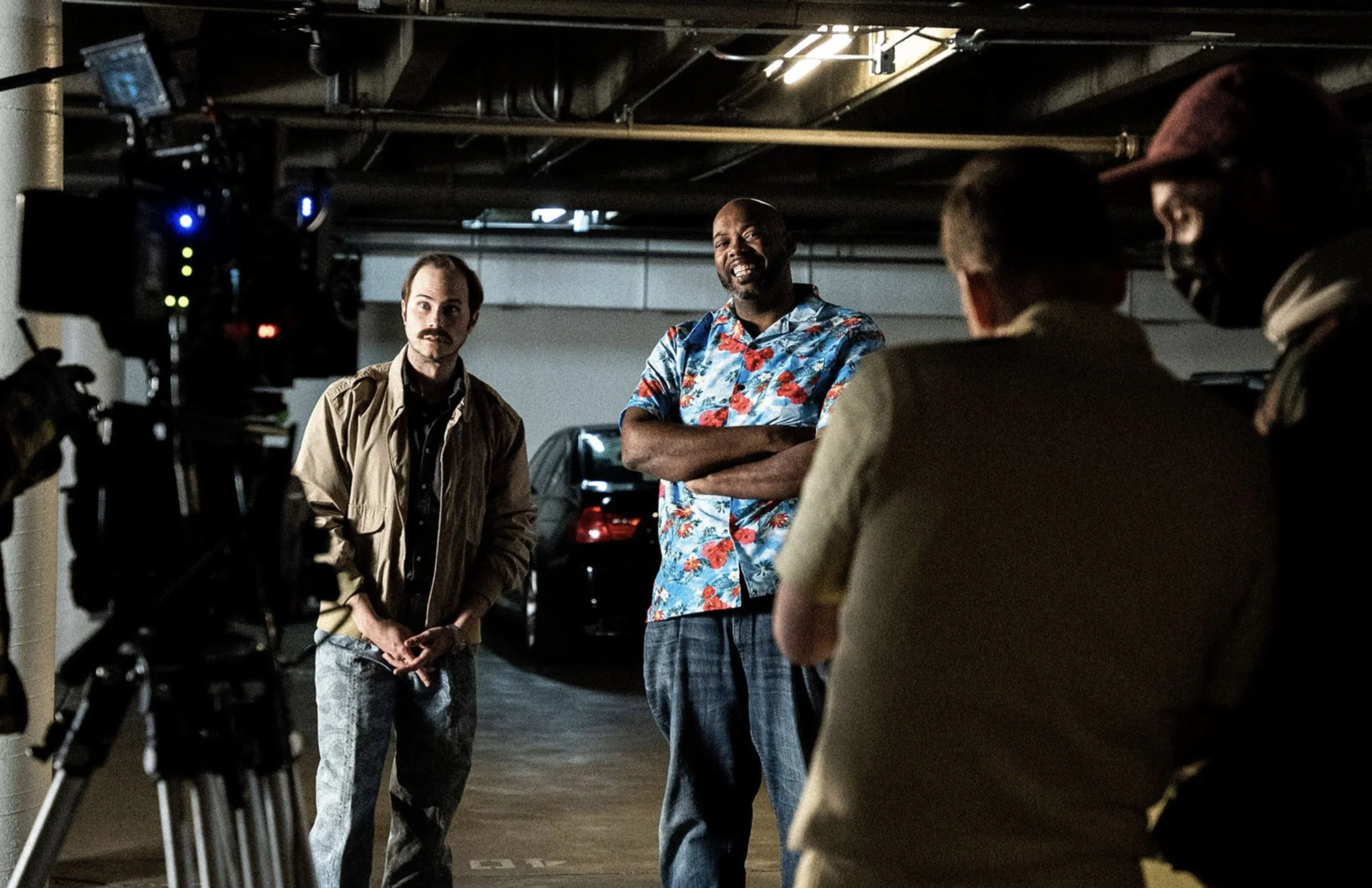 A group of men appear to be filming or discussing in a parking garage, with a camera on a tripod capturing the scene.