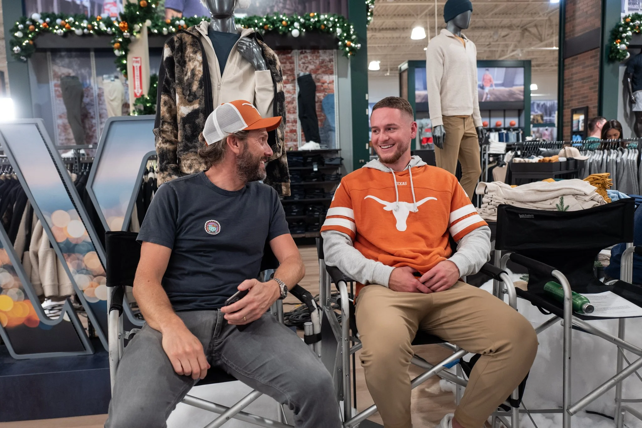 Two men sitting in folding chairs in a clothing store, smiling and talking to each other.