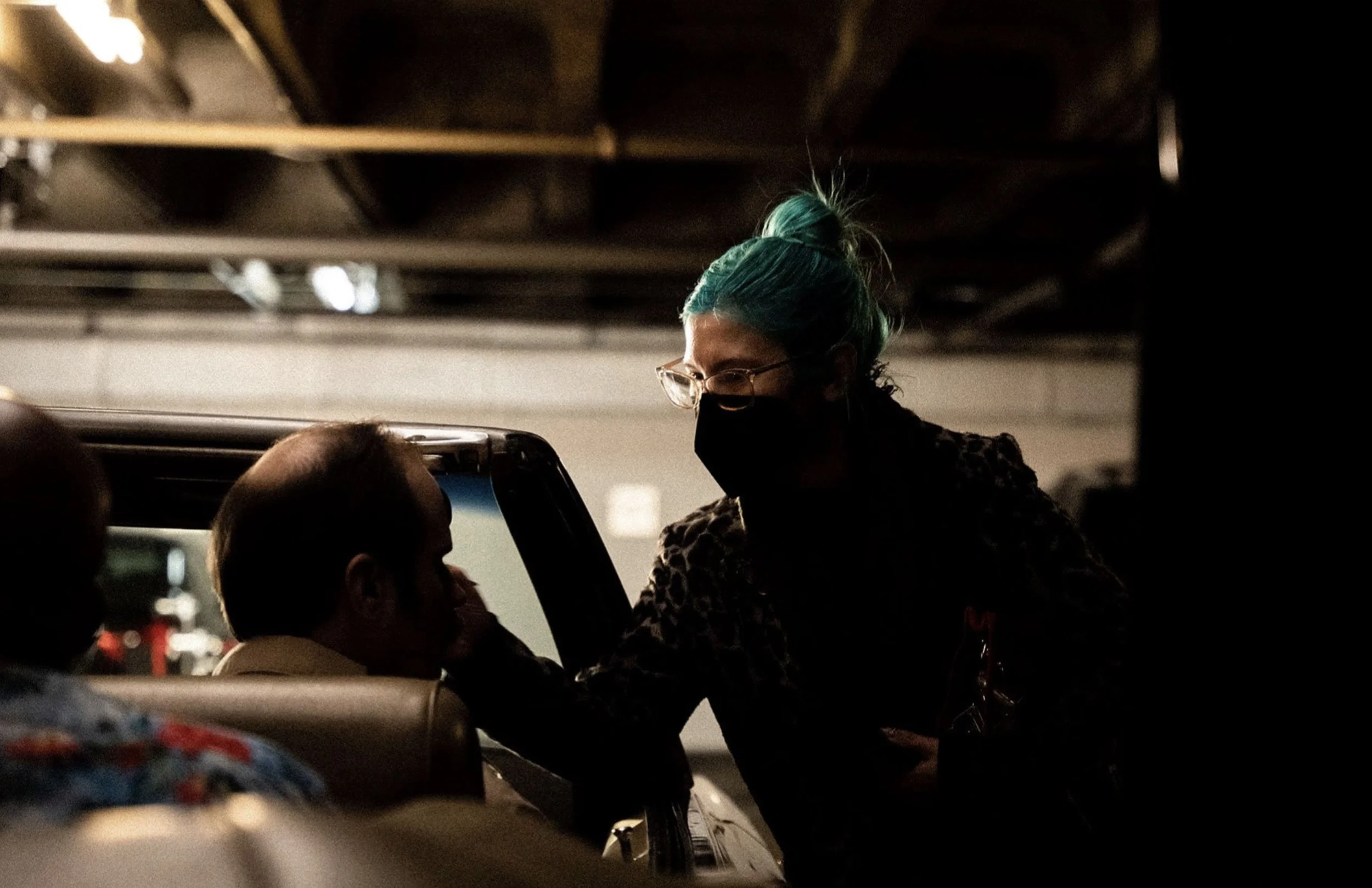 Person with teal hair and glasses leaning into car window, talking to man inside car, in a dimly lit underground parking garage, both wearing masks.