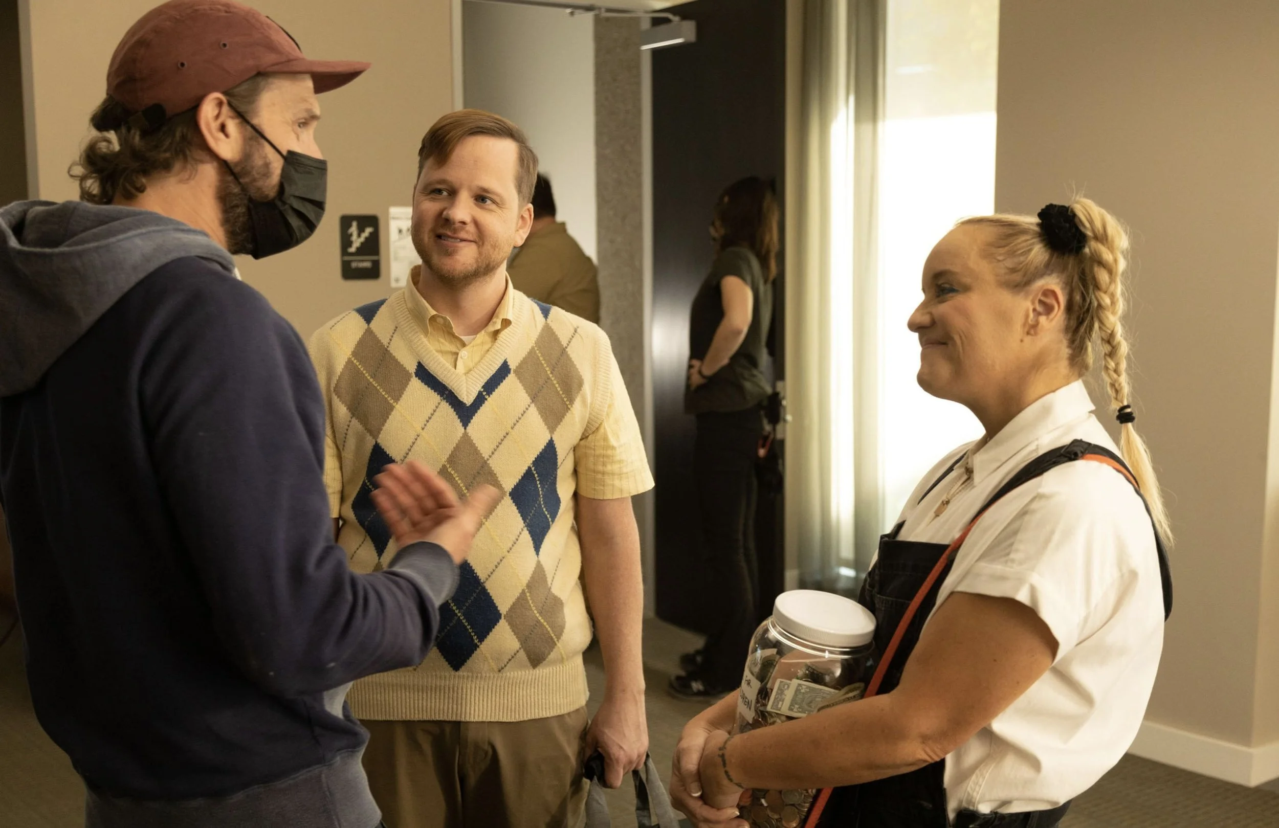 Three people engaged in conversation in an indoor setting, with a woman holding a jar filled with money and coins, two women standing in the background, and a man wearing a mask and a cap.
