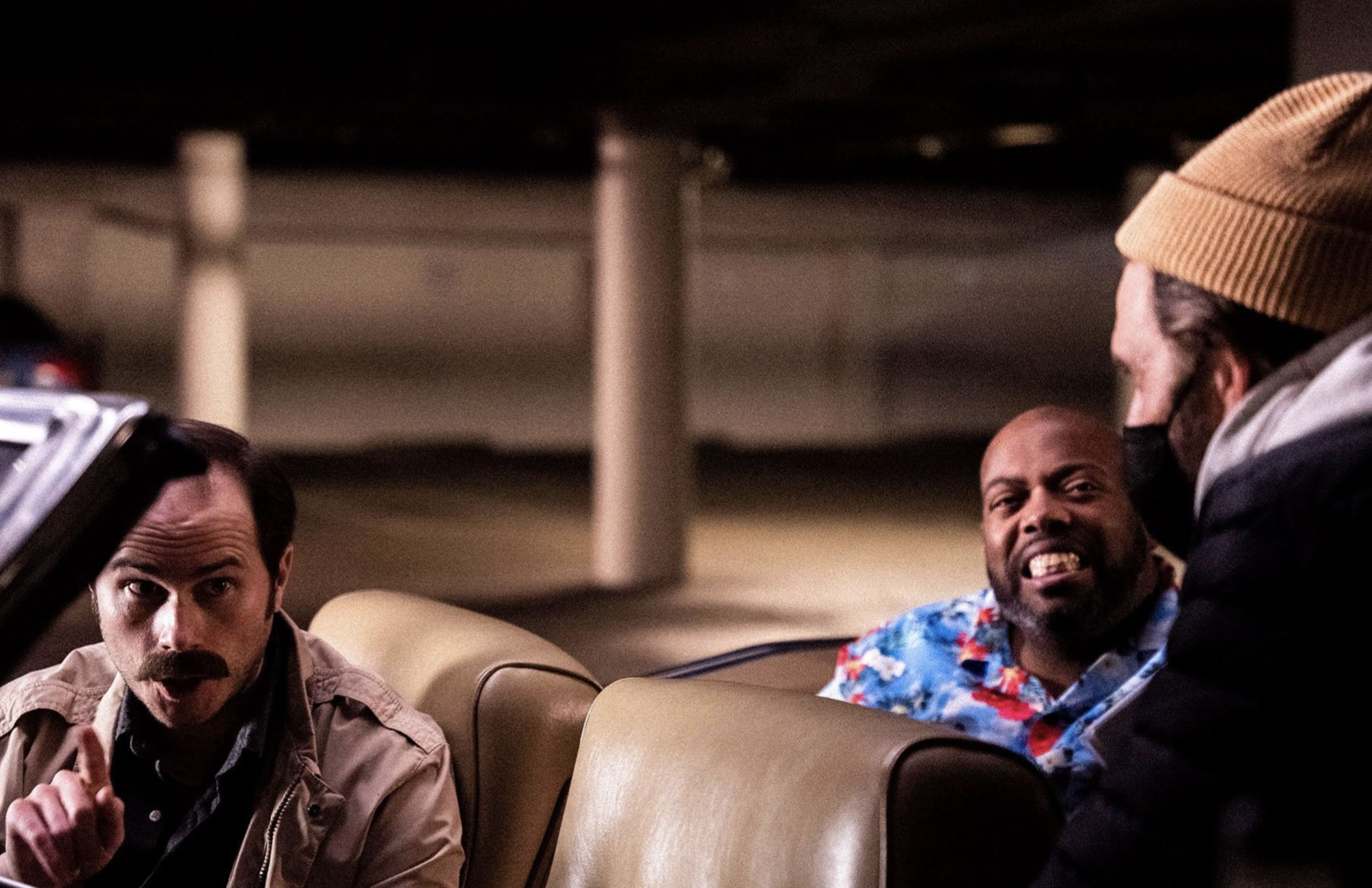 Three men sitting in a car, the man on the left has a serious expression with a mustache, the man in the middle has a big smile and is wearing a floral shirt, and the man on the right, wearing a beige beanie and face mask, is talking to the others.