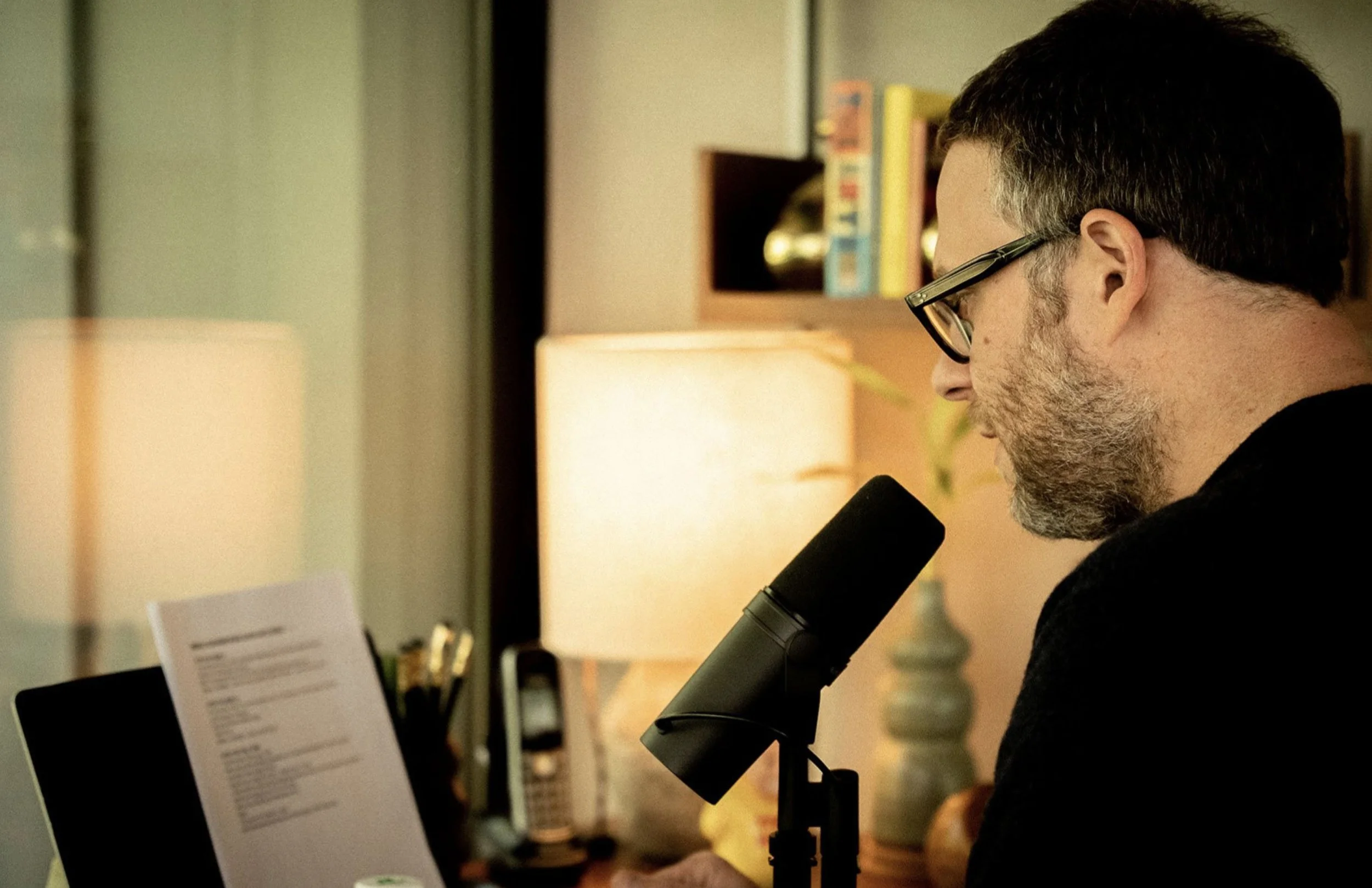 A man with glasses and a beard speaking into a microphone while reading a document in a cozy, well-lit room.