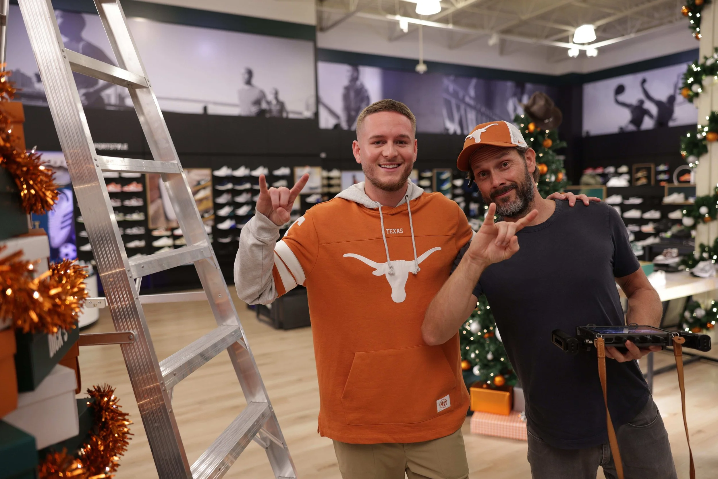 Two men smiling and posing inside a shoe store decorated for Christmas. One man is wearing an orange Texas Longhorns hoodie, and the other is wearing a black t-shirt and a black and orange Texas Longhorns cap. Christmas trees and holiday decorations 