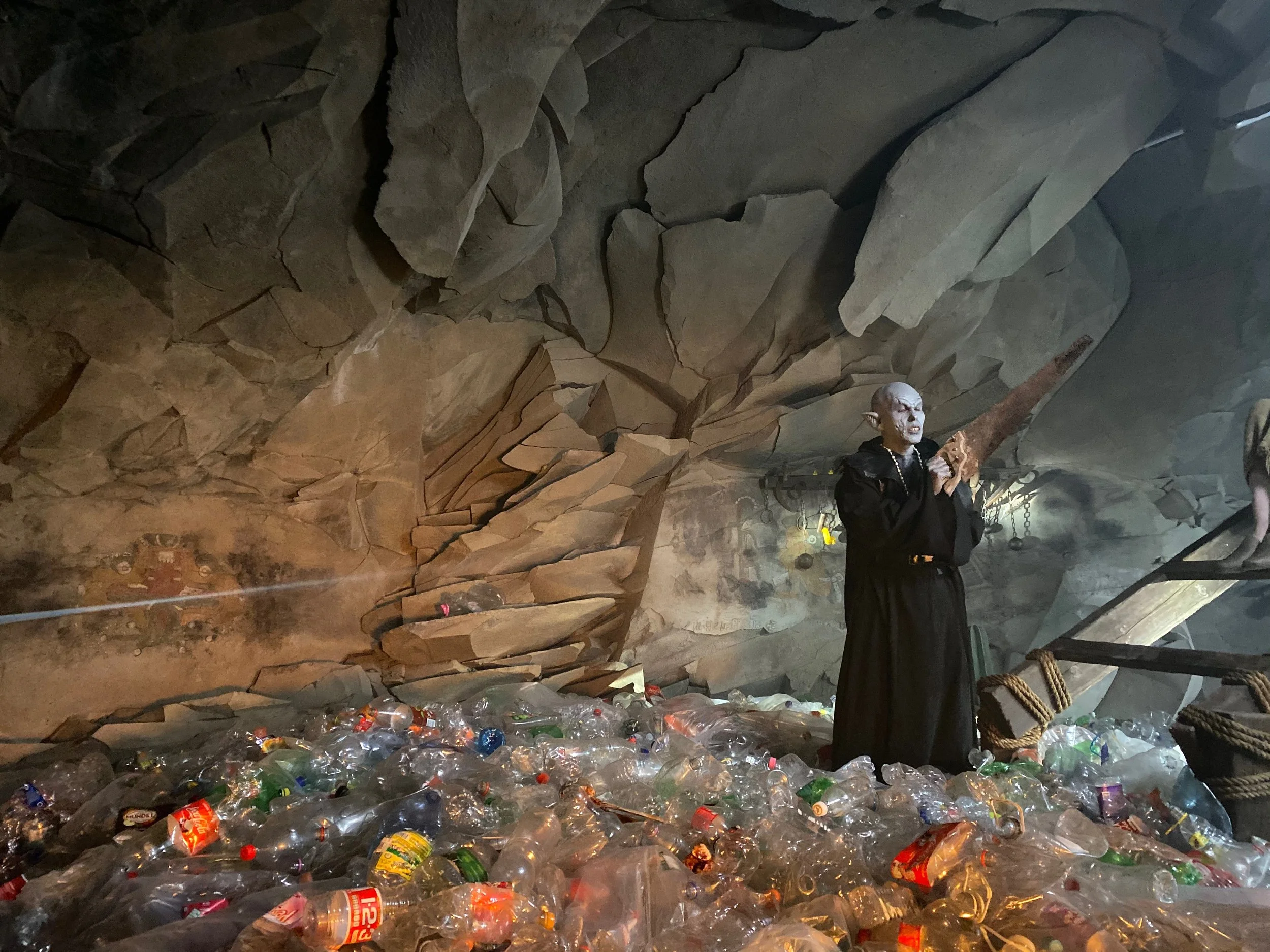 A person dressed as a goblin or goblin-like character holding a large axe in a cave-like environment filled with plastic bottles and trash.