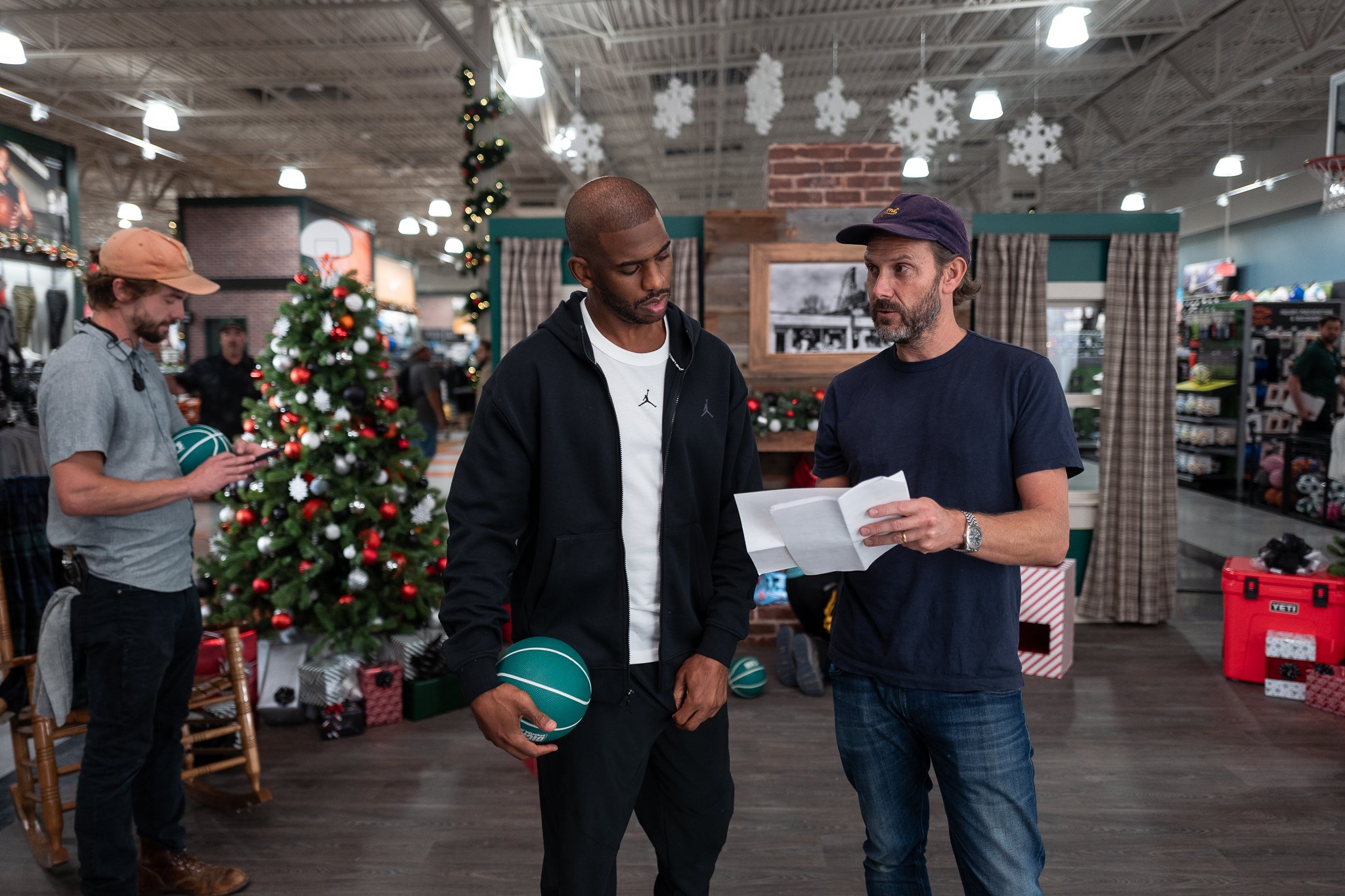 Two men having a conversation at a store decorated for Christmas. One is holding a basketball, and the other is holding a piece of paper. There is a Christmas tree and holiday decorations in the background.