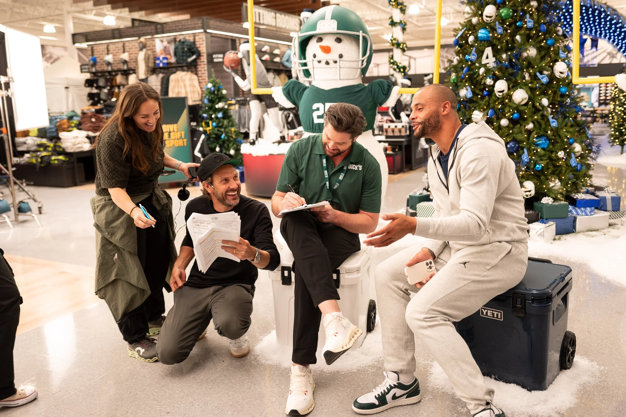 Group of five people in a store decorated with Christmas trees, one with a football helmet, having a lively discussion and laughing, with a snowman sports mascot in the background.