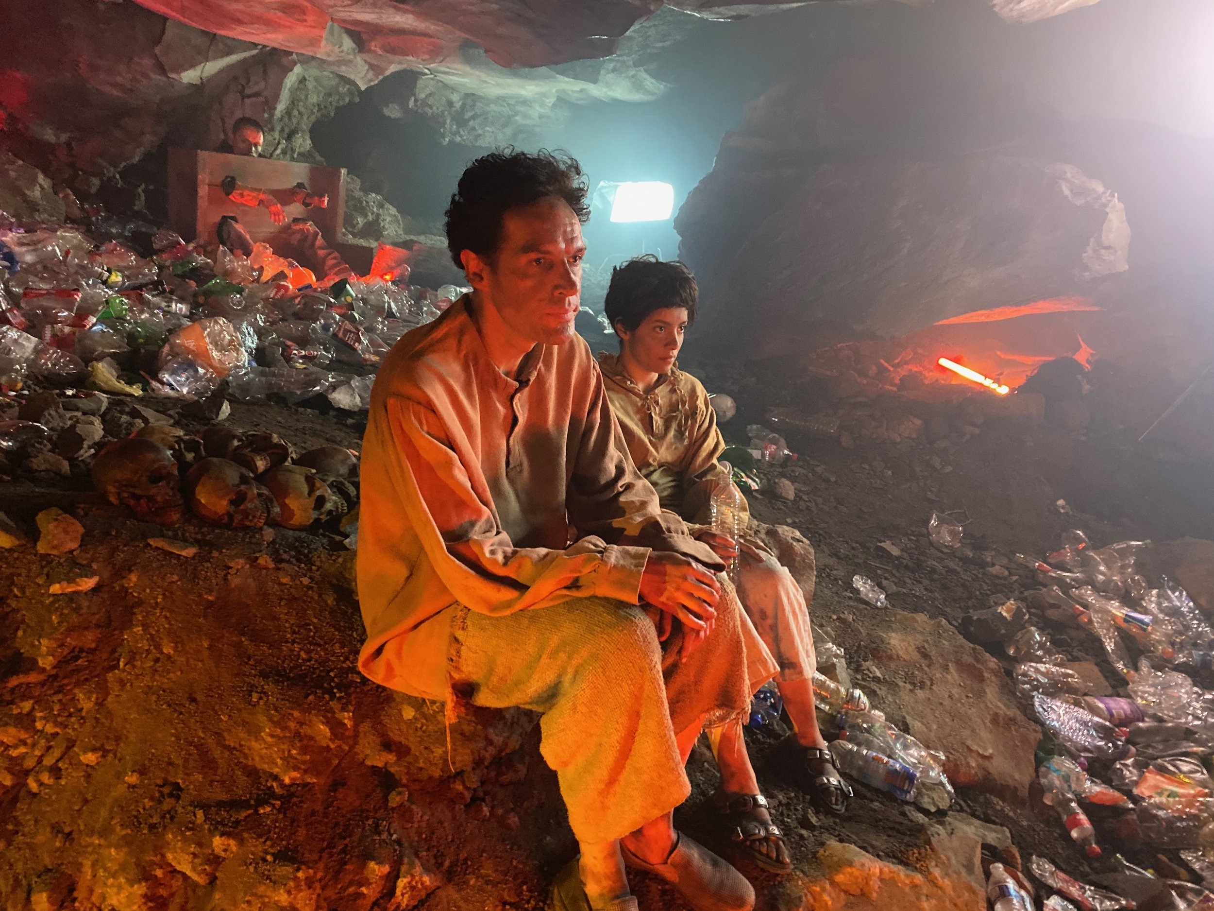 Two people sitting inside a cave surrounded by discarded plastic bottles and human skulls, with a faint red glow illuminating the darkness.