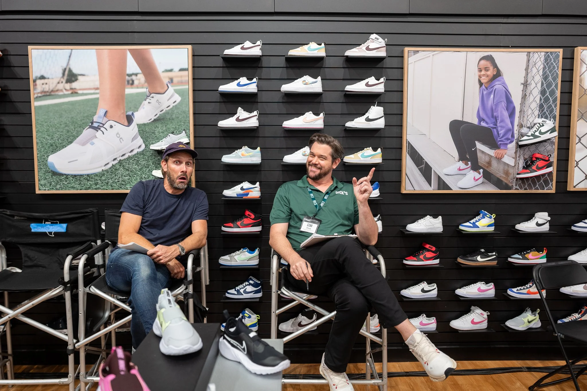 Two men sitting in chairs surrounded by sneaker display in store, one speaking and smiling, other looking surprised, with large sneaker photos on wall behind them.