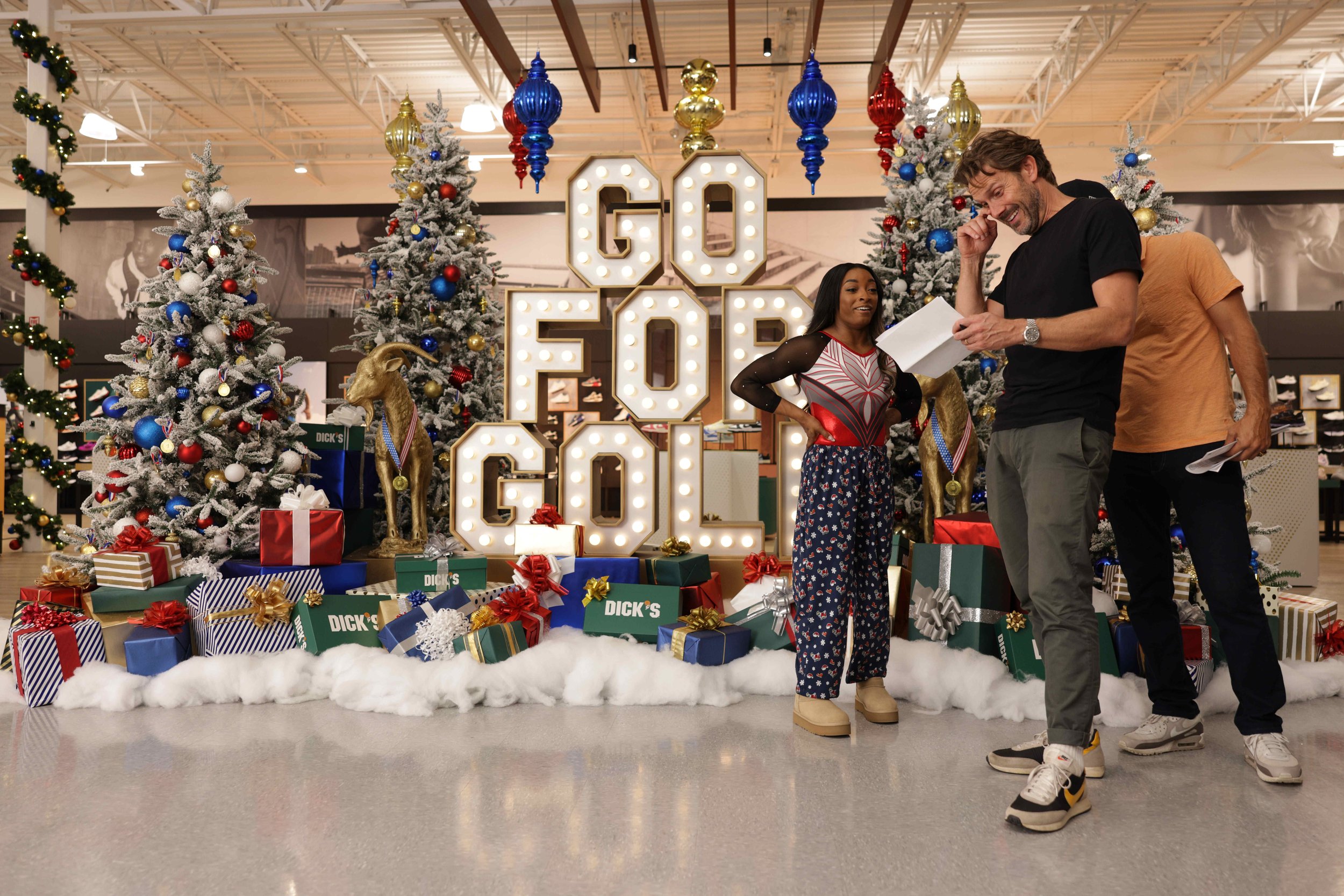 People shopping at a Christmas display with decorated trees, presents, and a large illuminated 'GOLD G' sign in the background.