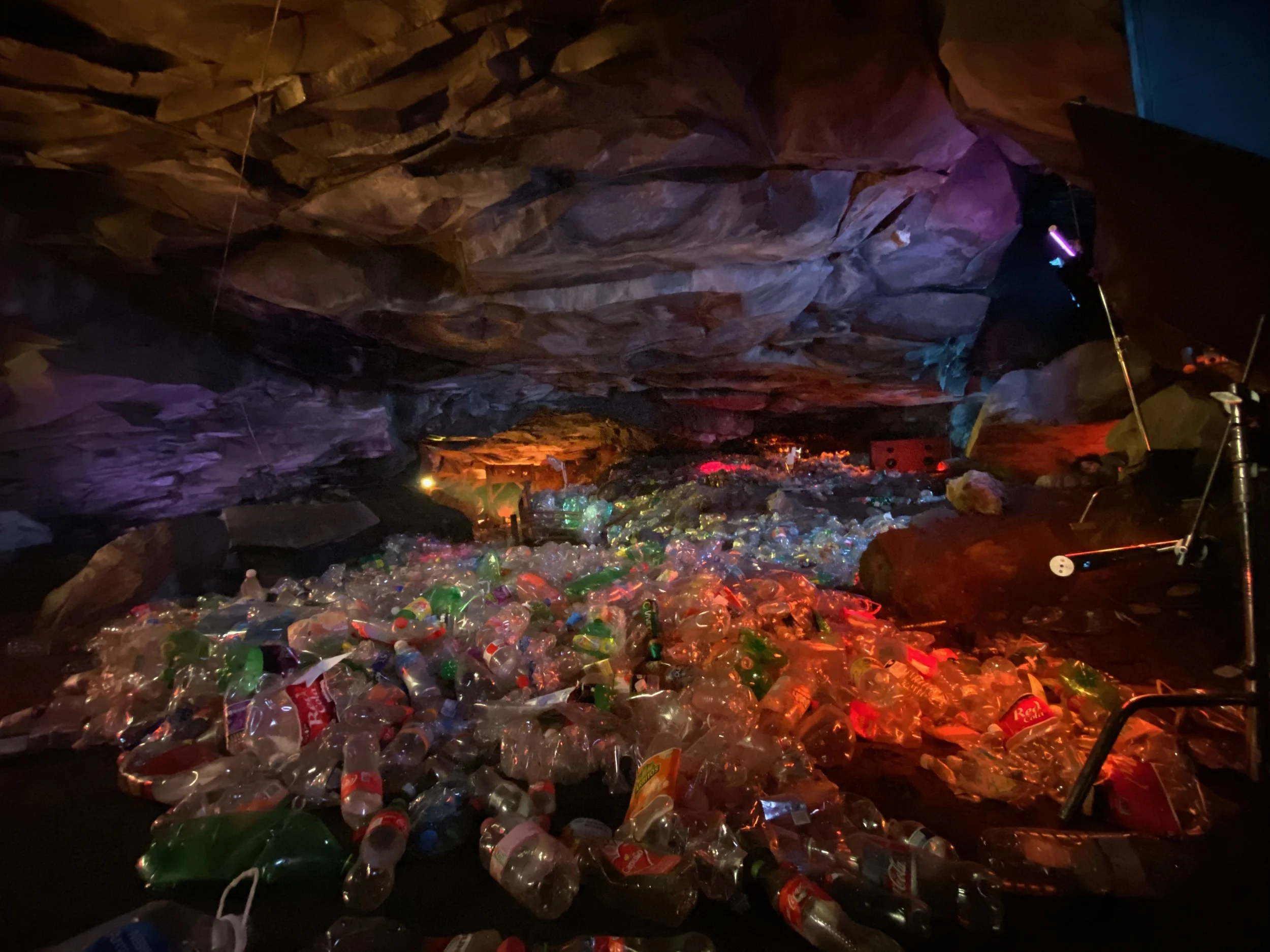 A cave filled with a large amount of plastic bottles and trash, illuminated by colorful artificial lights.