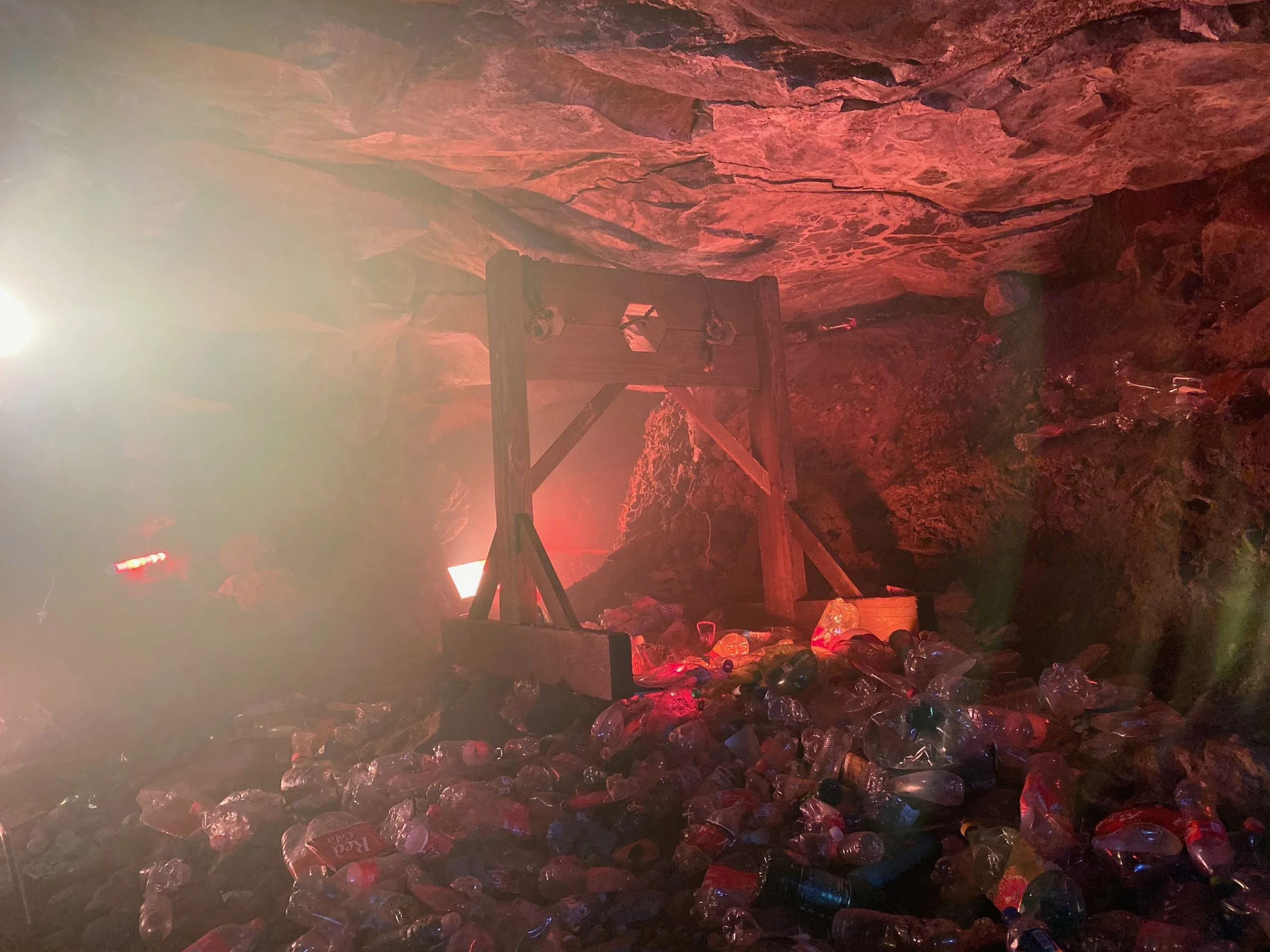 Inside a cave filled with plastic bottles and trash, with a wooden structure and a faint light source creating a hazy atmosphere.