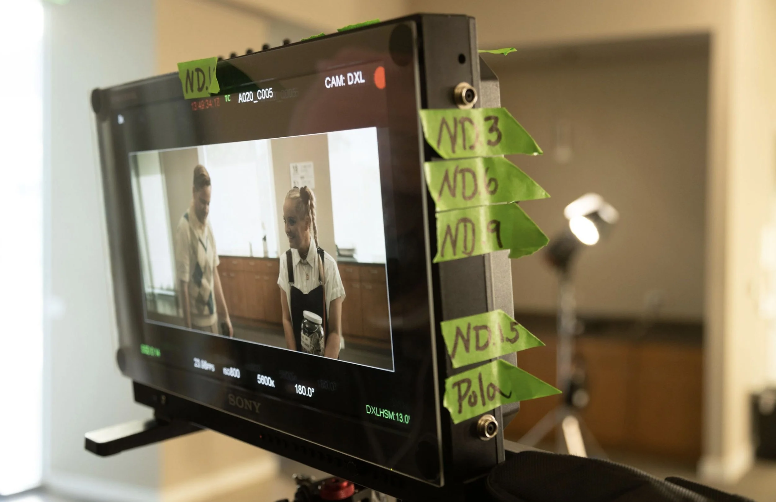 Scene from a film or video shoot showing a monitor displaying two young women, one in a white shirt with a black vest and the other in a similar outfit, in a room with wood cabinets and large windows. The monitor is marked with color correction and l