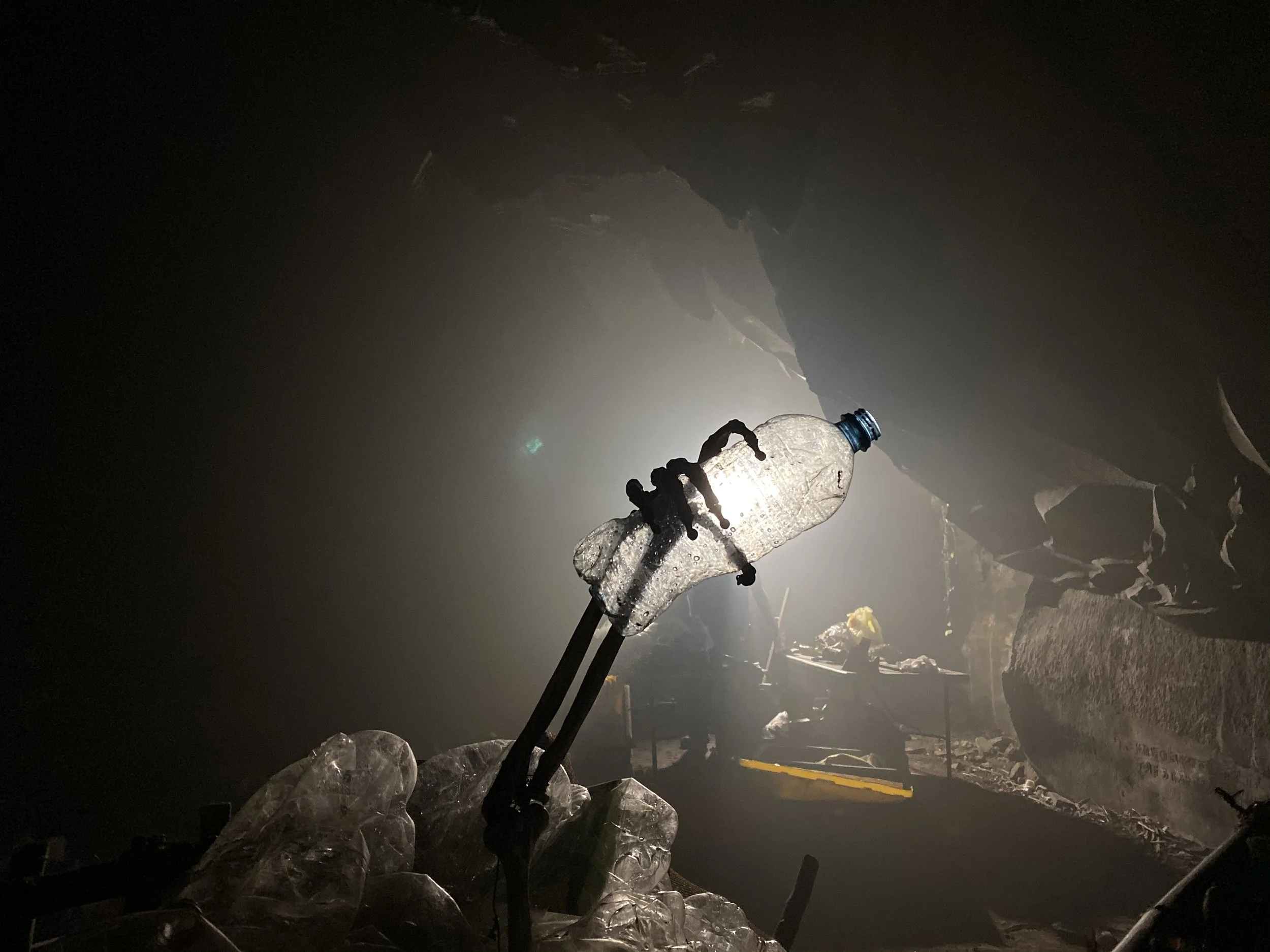A plastic water bottle suspended by black cables, illuminated by bright light inside a dark, cave-like environment with rocks and debris.