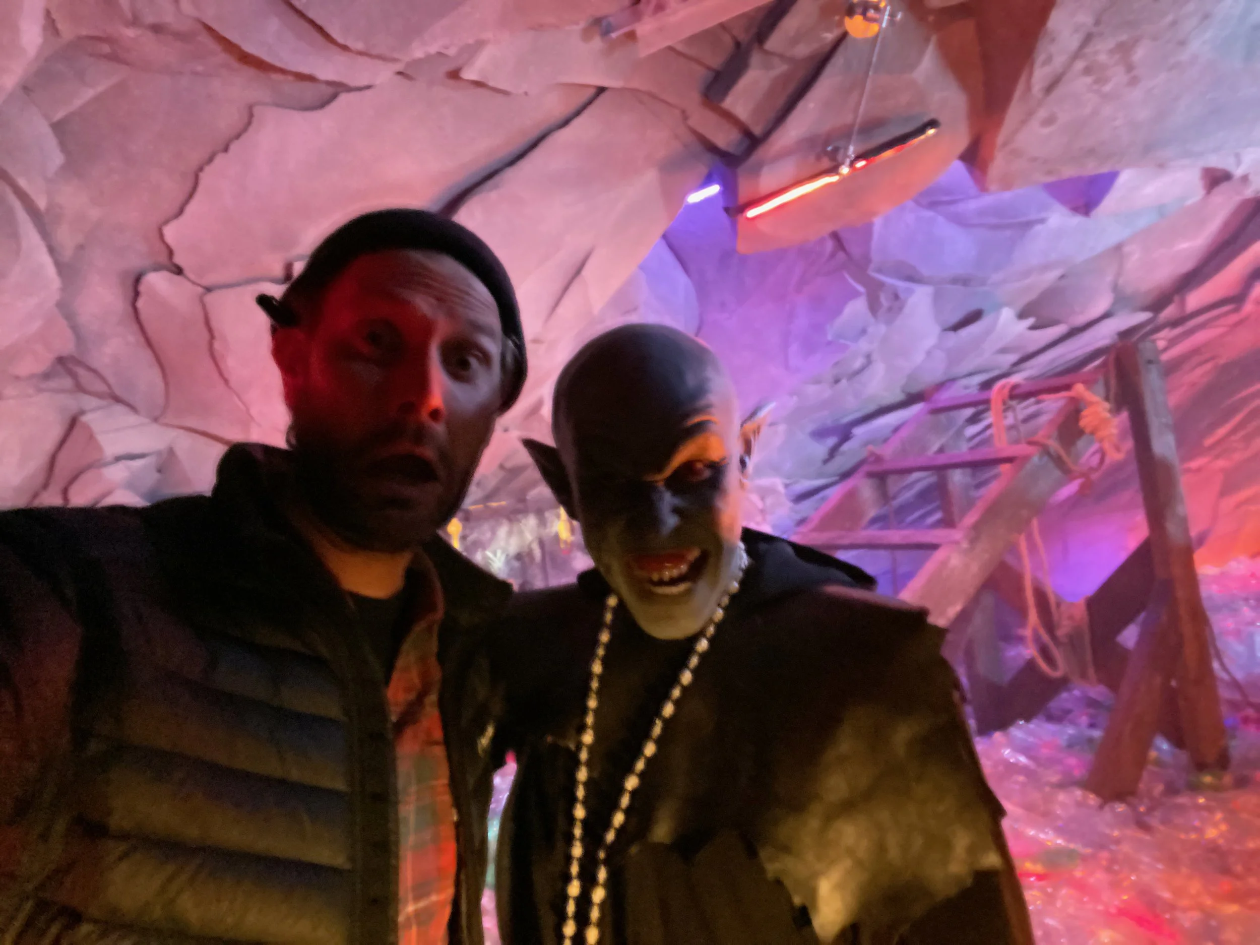 A person taking a selfie with a person in a costume resembling a fantasy character in a cave with rock walls and colorful lighting.