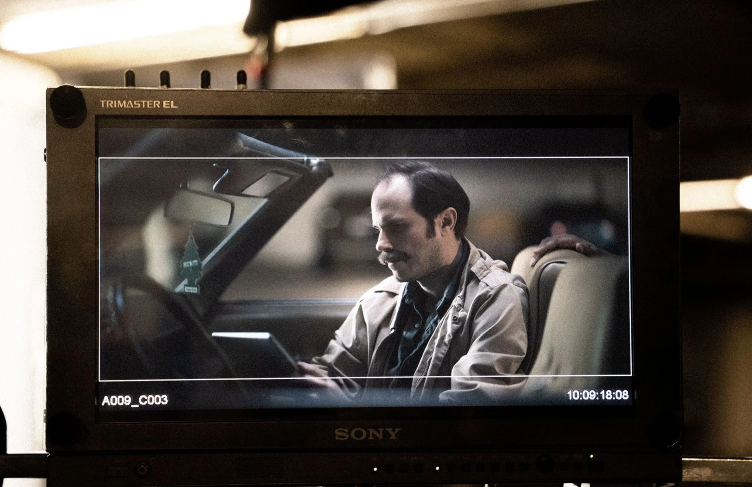 A man with dark hair, mustache, and beard sitting in a vintage beige car, looking at his phone while the hand of another person rests on his shoulder.