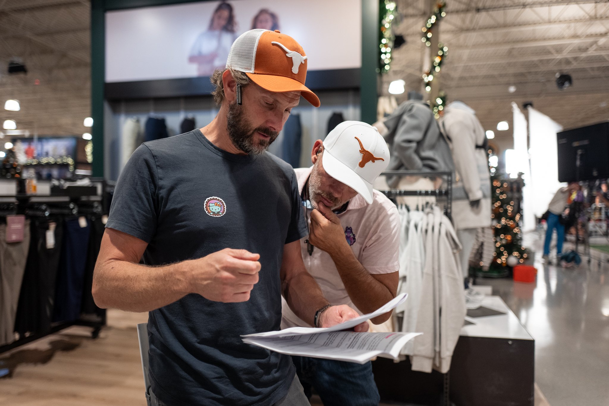 Two men shopping indoors, wearing Texas Longhorns caps, examining documents or brochures, with clothing displays and Christmas decorations in the background.