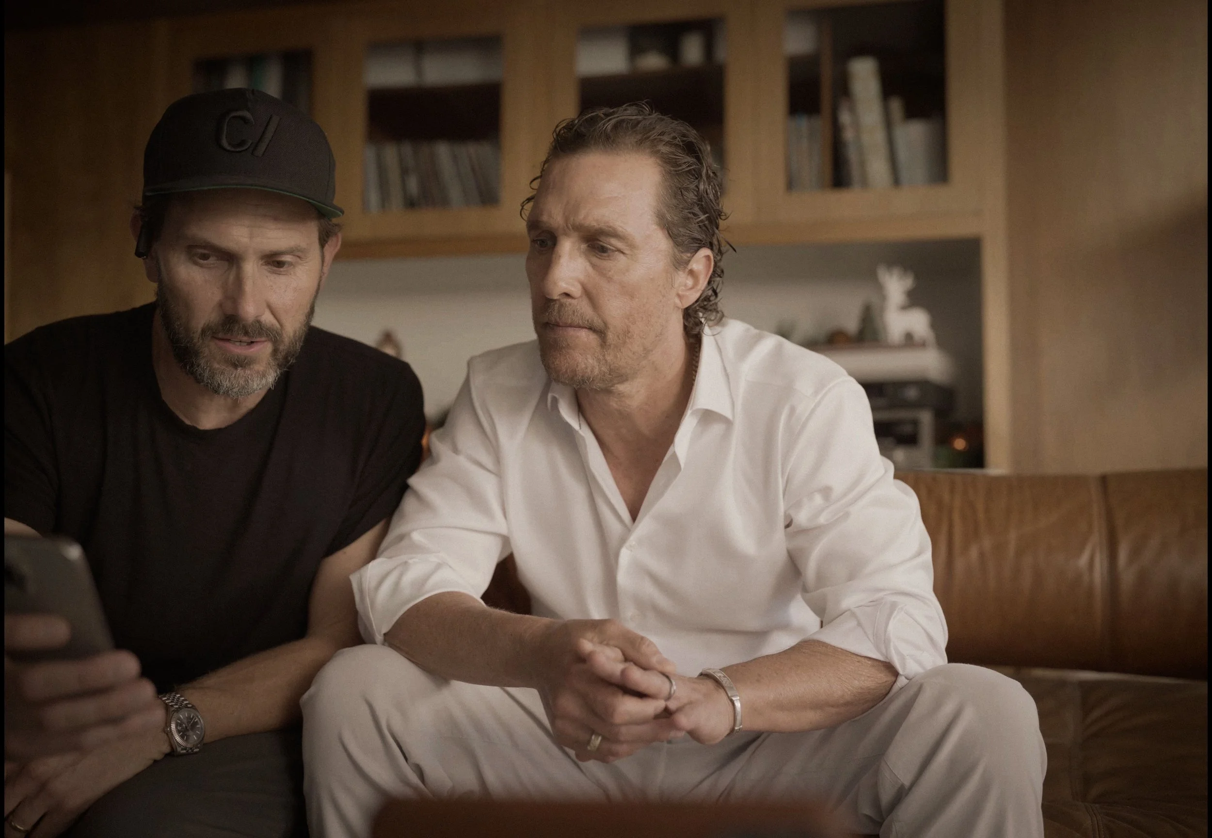 Two men sitting on a brown leather couch looking at a phone. The man on the left is wearing a black t-shirt, a black cap, and a wristwatch. The man on the right is dressed in a white shirt and beige pants with hands clasped. Background includes woode