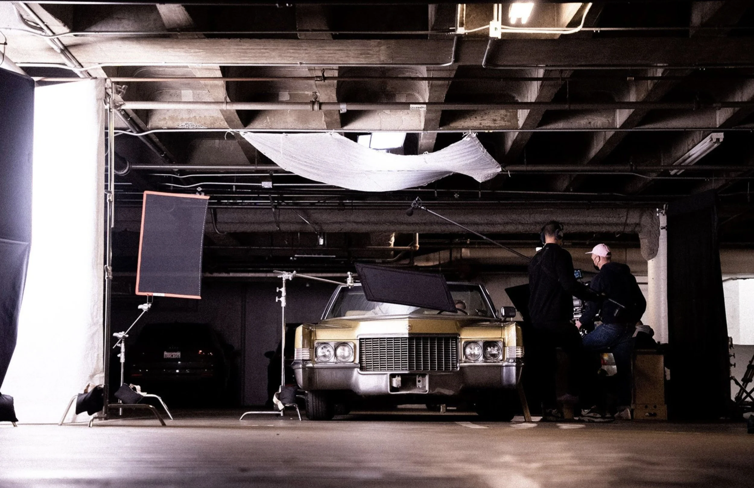 Film set with a vintage car in the center, surrounded by lighting equipment, crew members working, and a partially covered ceiling in an industrial space.