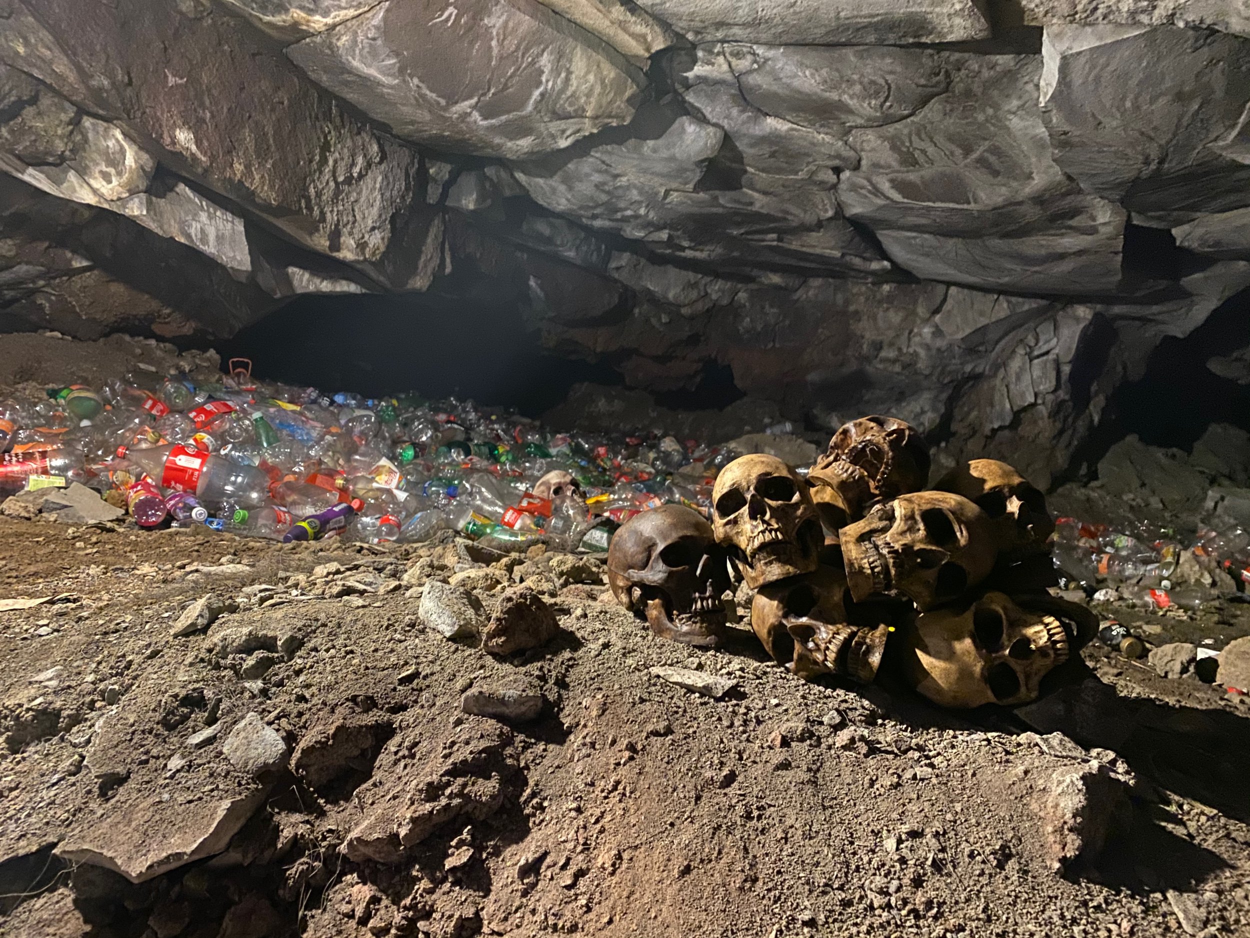 A pile of human skulls in a dark cave with trash, including plastic bottles and cans, spread across the rocky ground.