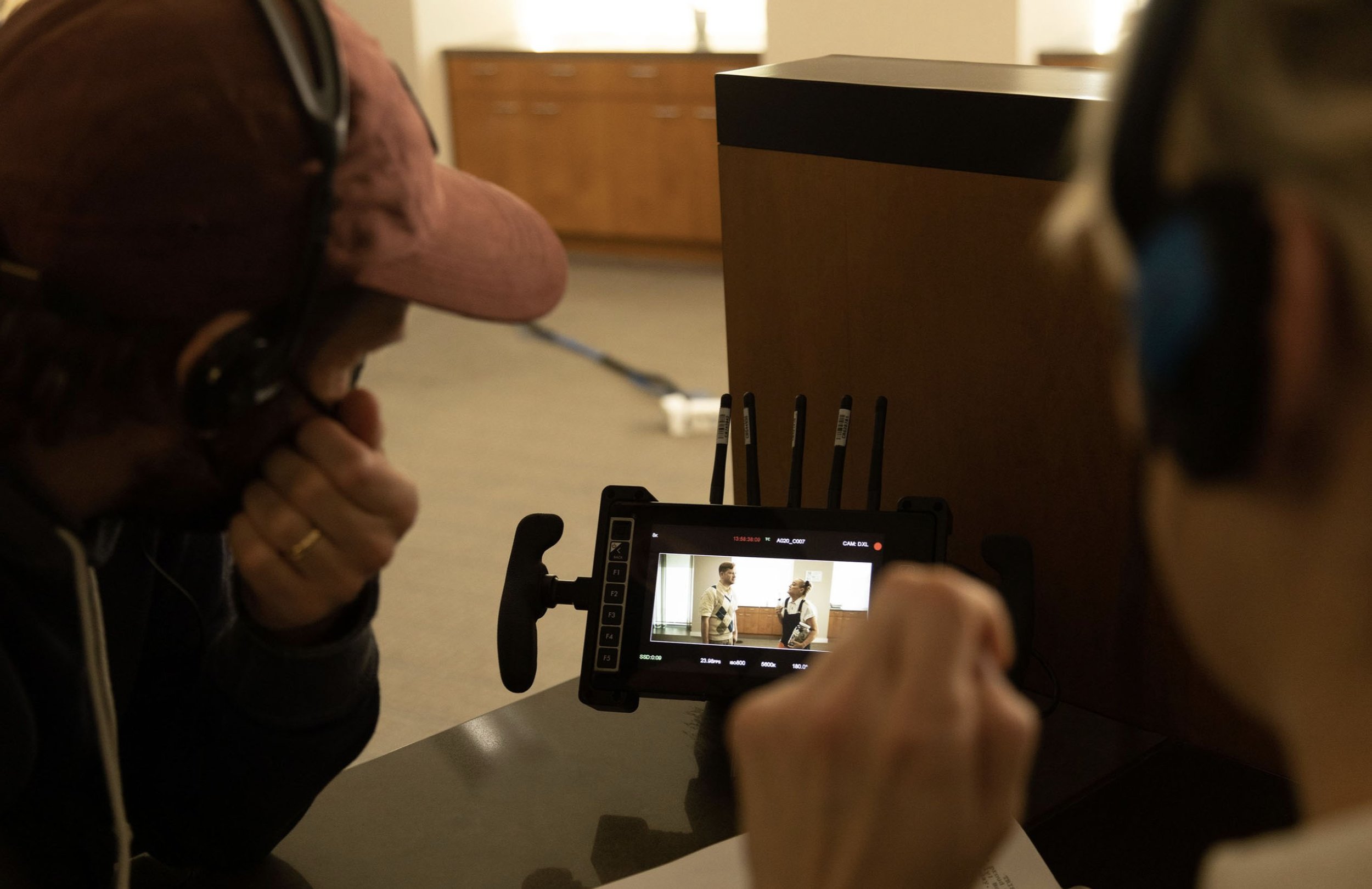 Two people on a film set, one with a pink hat and headset, the other with a blue headset, watching a monitor showing them in front of a wooden background in a room with beige carpeted floor.