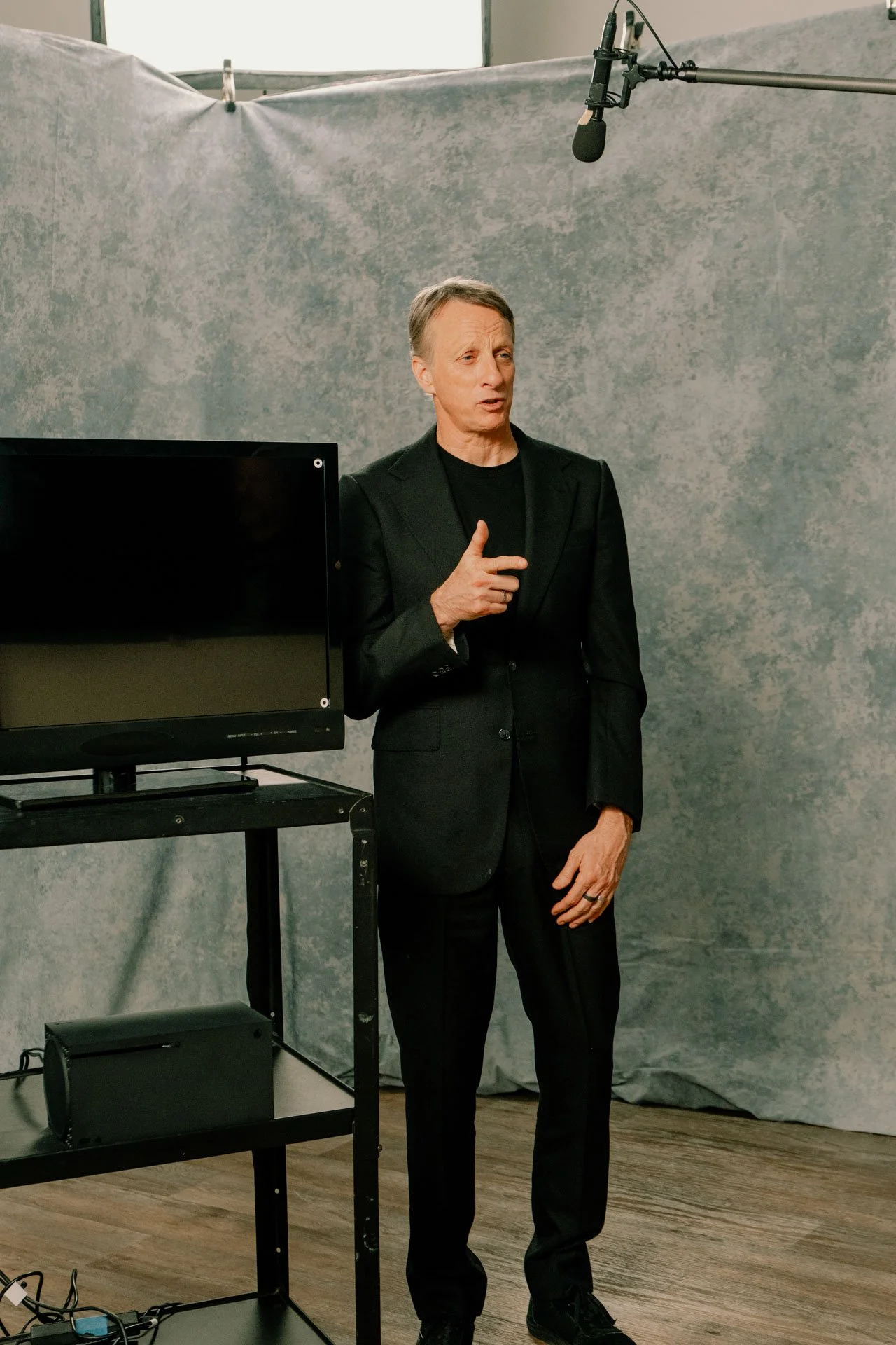 A woman in a black suit stands in front of a gray background with a TV and a microphone overhead, giving a speech or presentation.