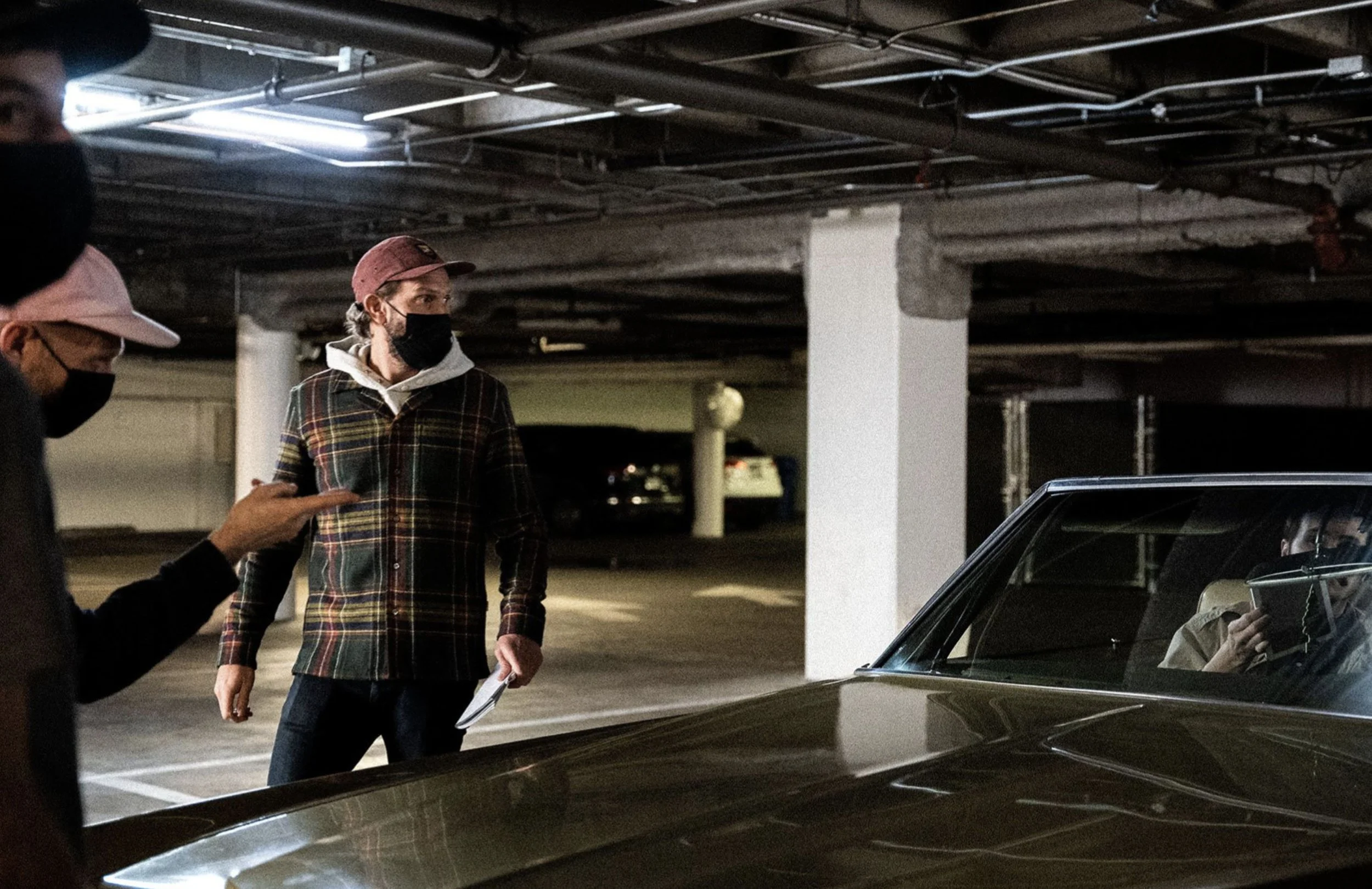 Three people in a parking garage, two of them standing and one sitting inside a vintage car, all wearing masks. The person inside the car is holding a tablet. The garage has exposed pipes on the ceiling and white columns.