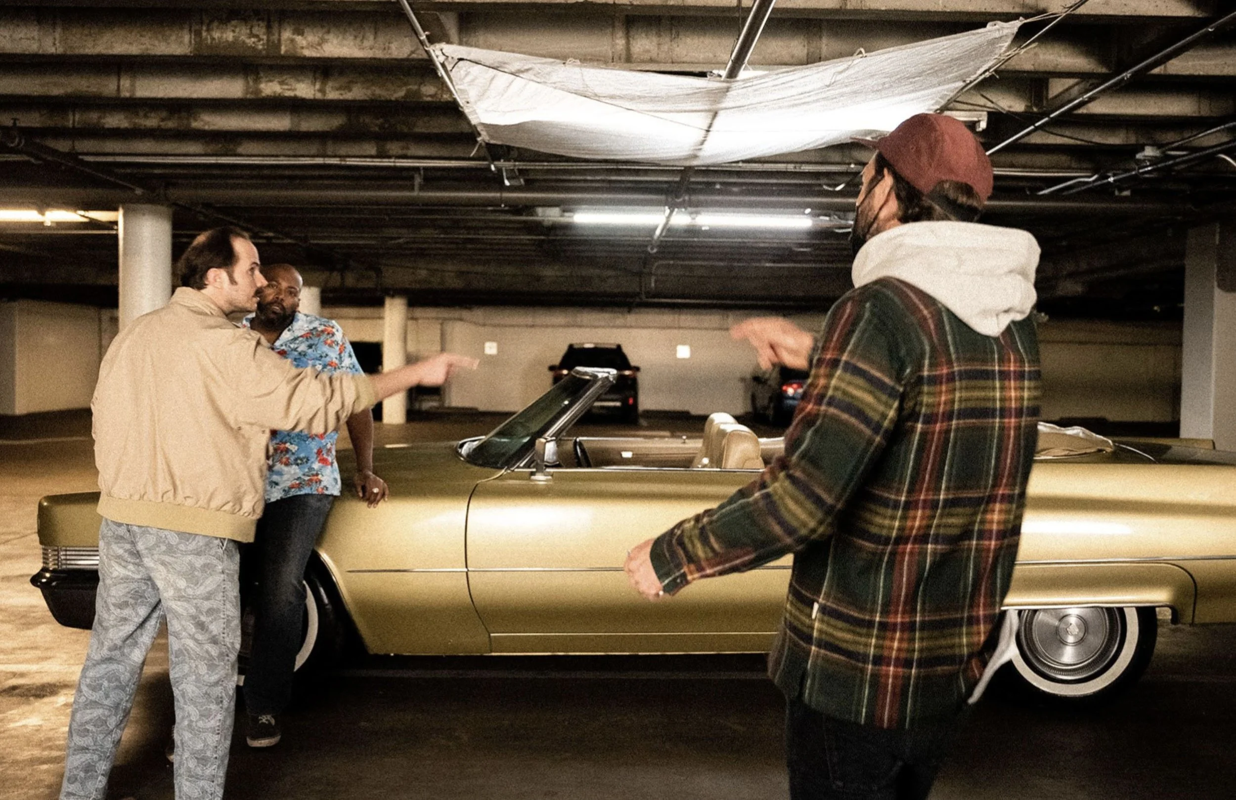Three men having a discussion in a parking garage, standing next to a vintage gold convertible car.