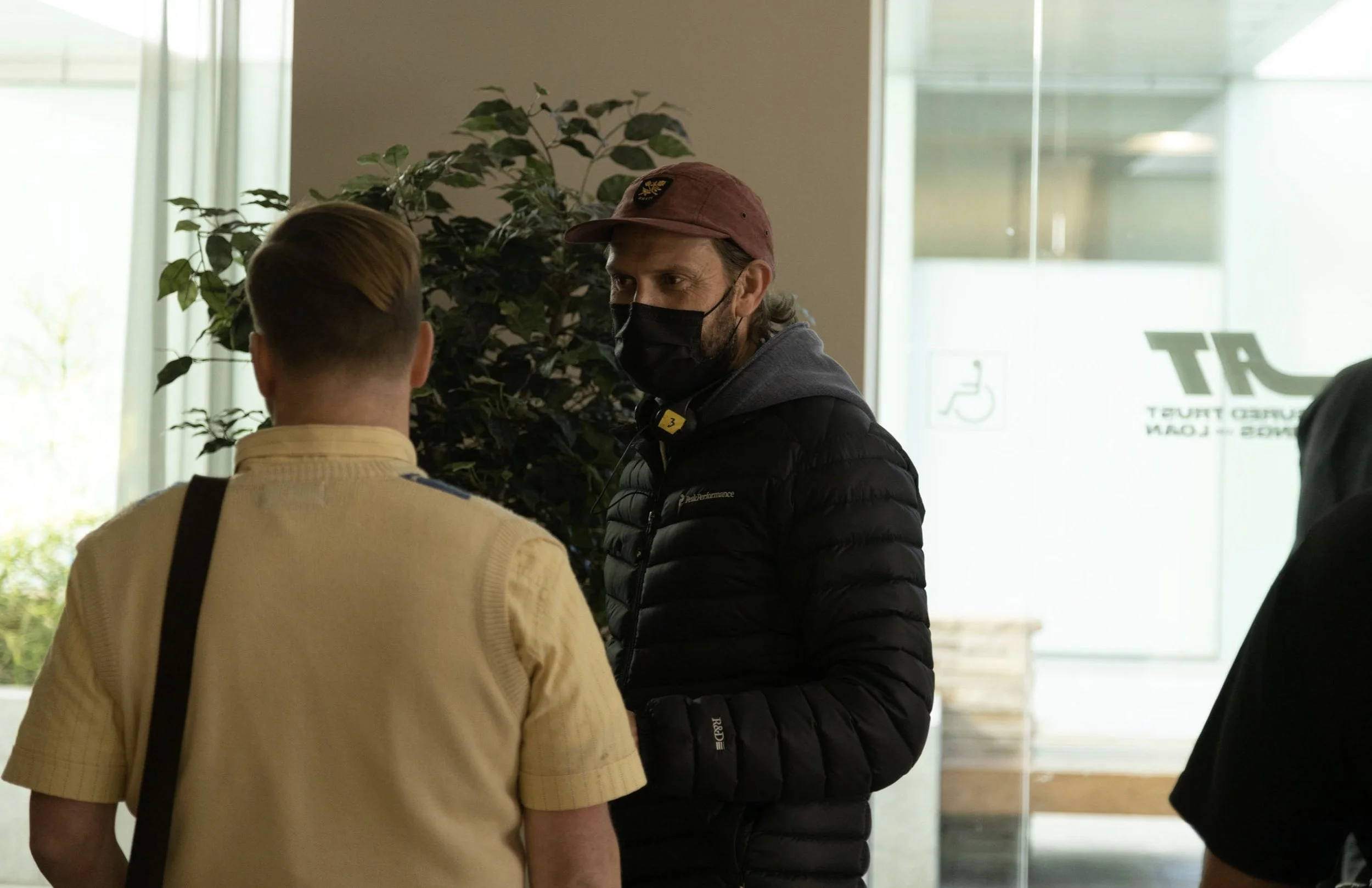 Two men engaged in conversation inside a building near a large window with natural light. One man is wearing a yellow shirt and a backpack, while the other is wearing a black jacket, a cap, and a face mask.