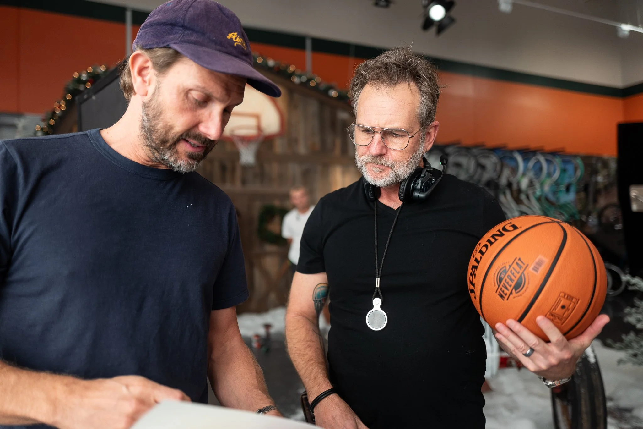 Two men, one with a baseball cap and the other with glasses, stand together in a gym or sports store, looking at a paper or device. The man with glasses holds a basketball.