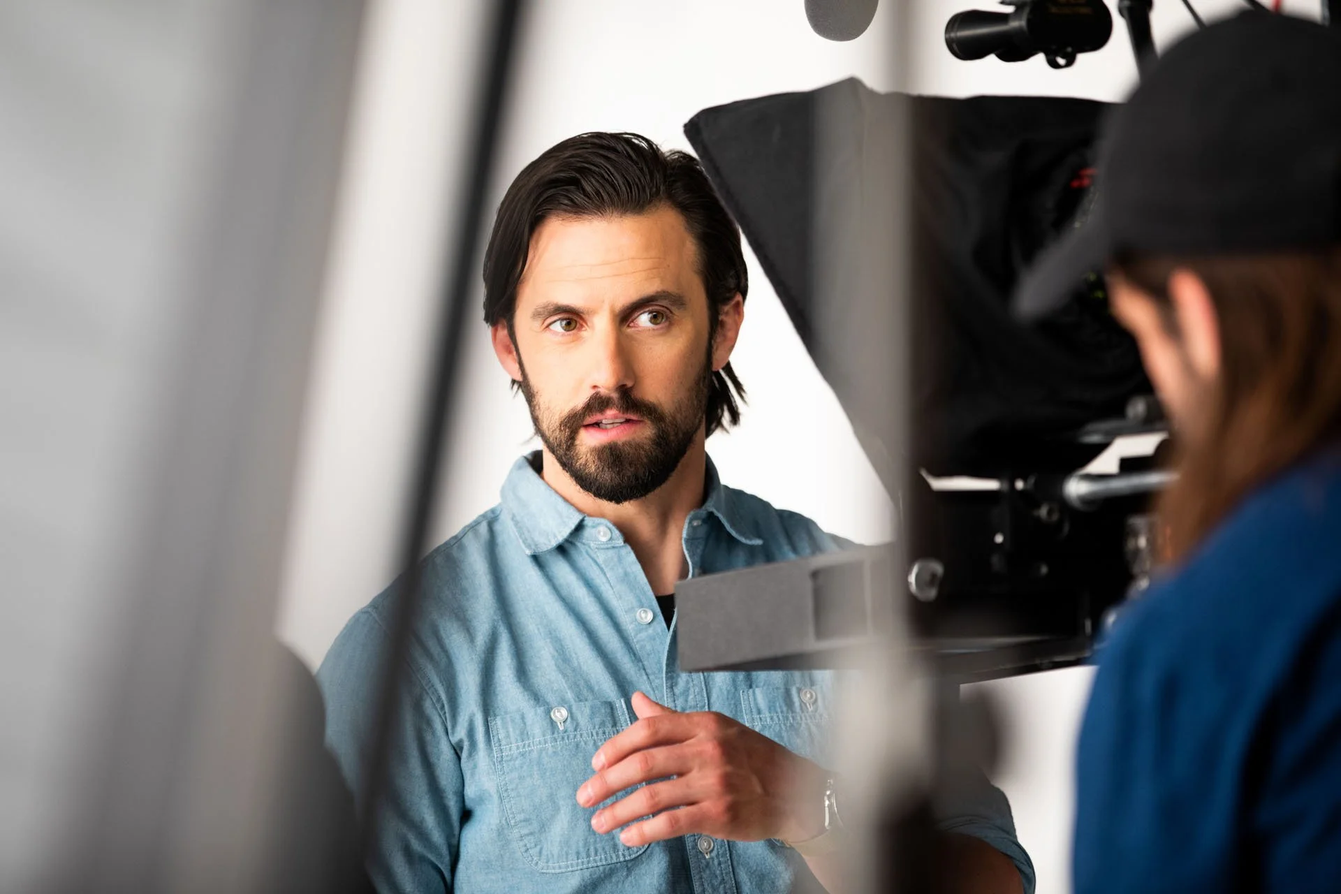 A man with dark hair and a beard, wearing a light blue denim shirt, looks thoughtfully while being filmed or photographed, with a camera and a person in a black hat nearby.