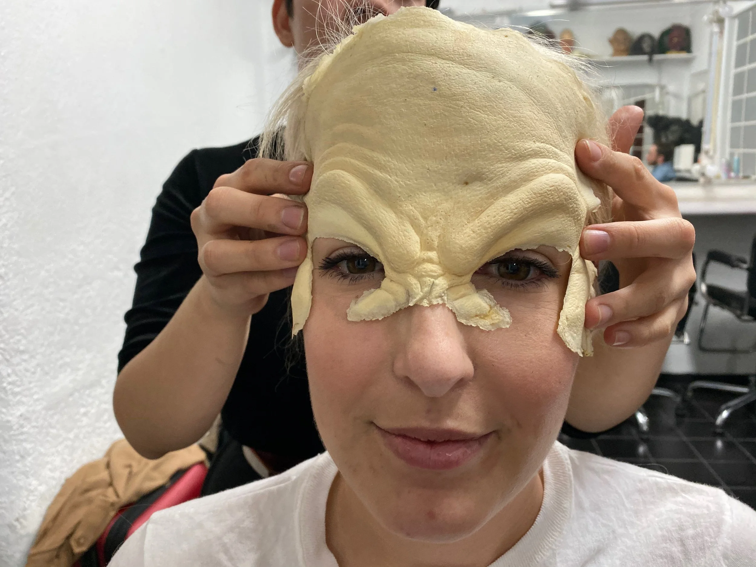 A woman having a skate mask applied to her face by another person in a room with shelves and chairs in the background.