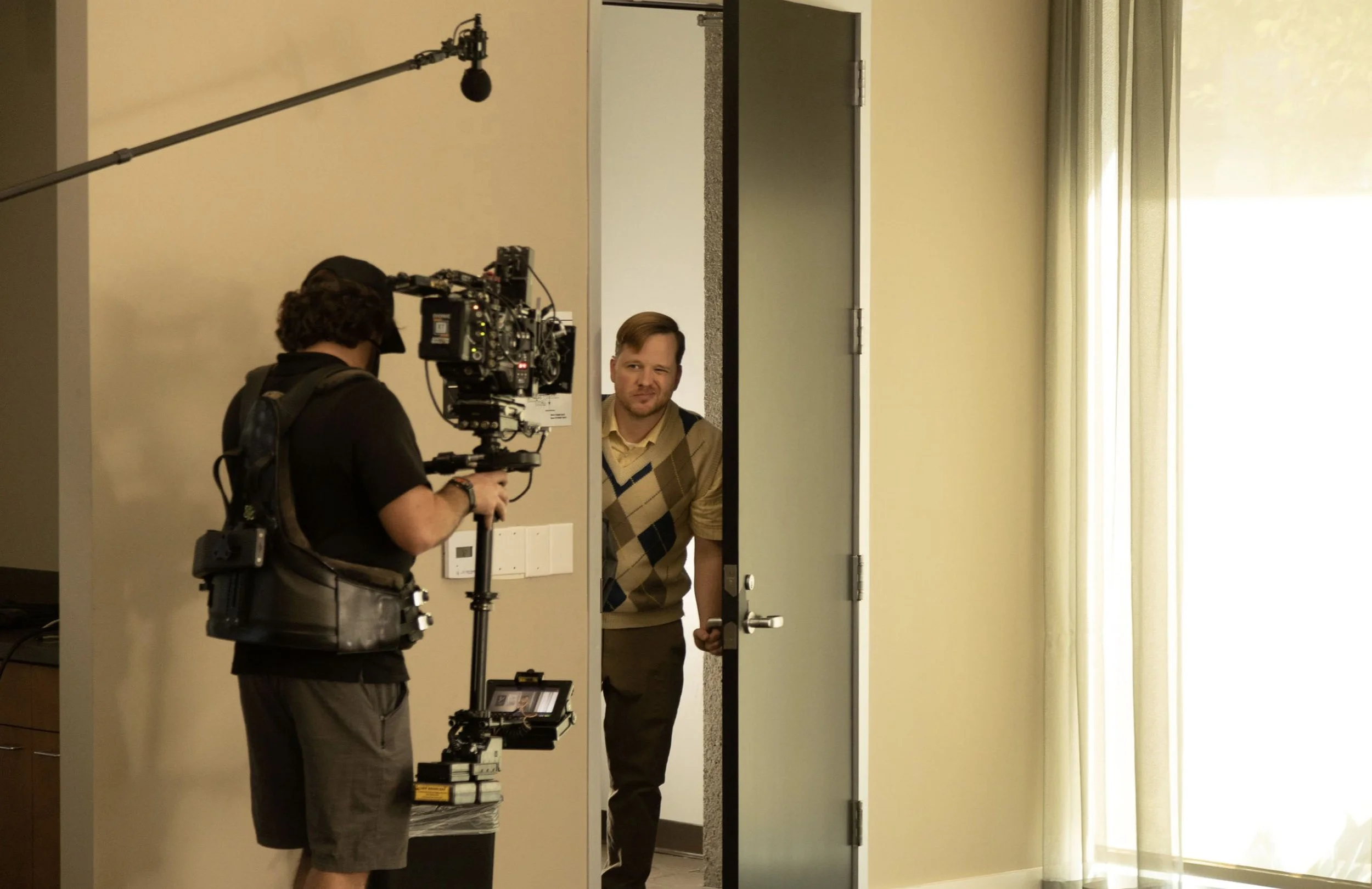 A man wearing a beige argyle sweater vest and khakis is peeking out from behind a partially open door, filming a man inside a room with a professional camera mounted on a stabilizer rig. The room has beige walls, a window with curtains to the right, 