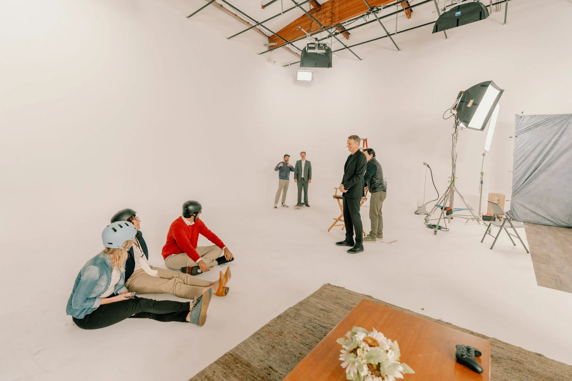 People on a photo shoot with a blank white backdrop, some are sitting on the ground wearing helmets, others are standing or working near lighting equipment and a director's chair.