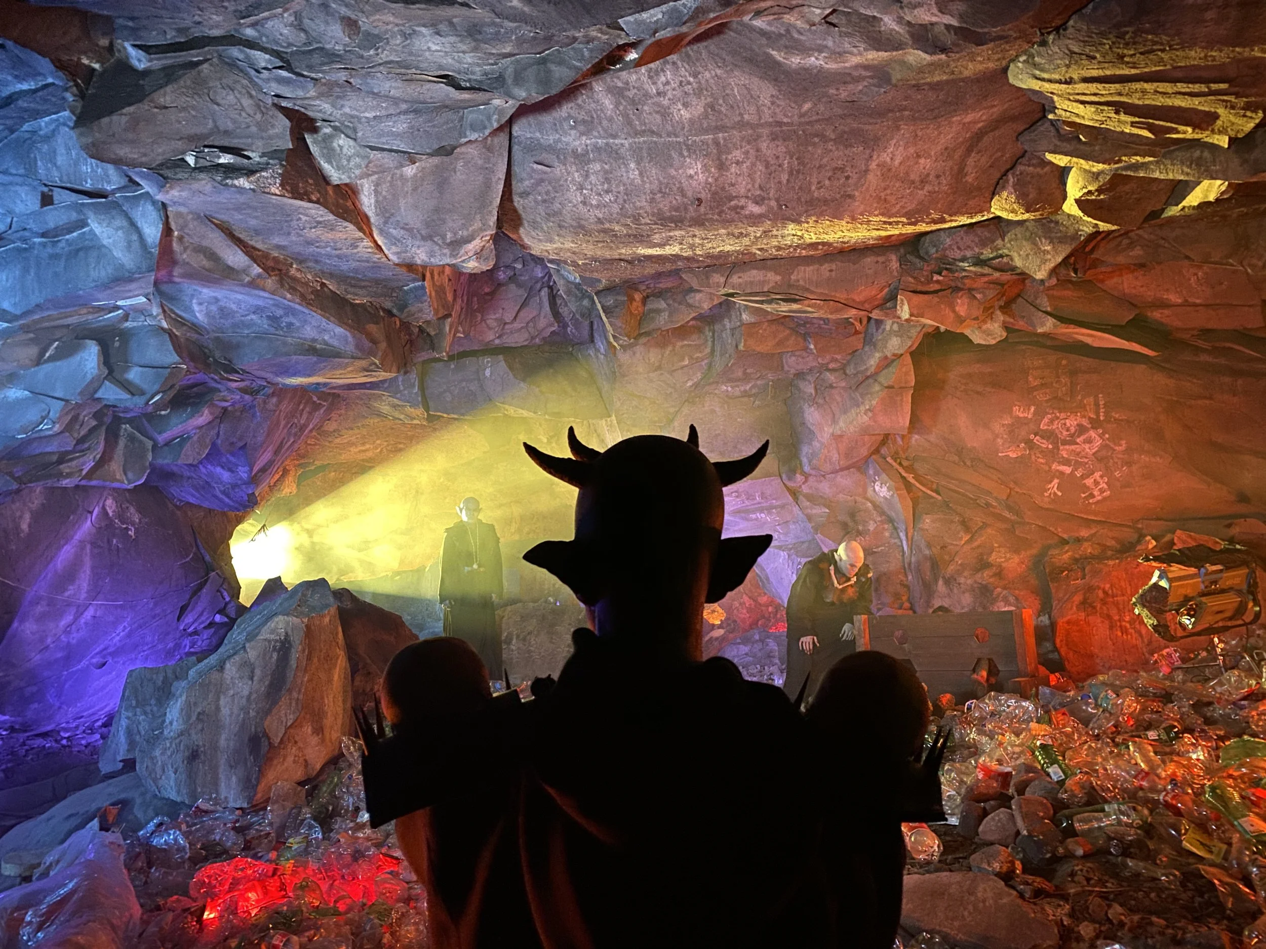 Person dressed as a devil with horns and pointy ears watches a performance in a colorful, rocky underground space filled with trash and colored lights.