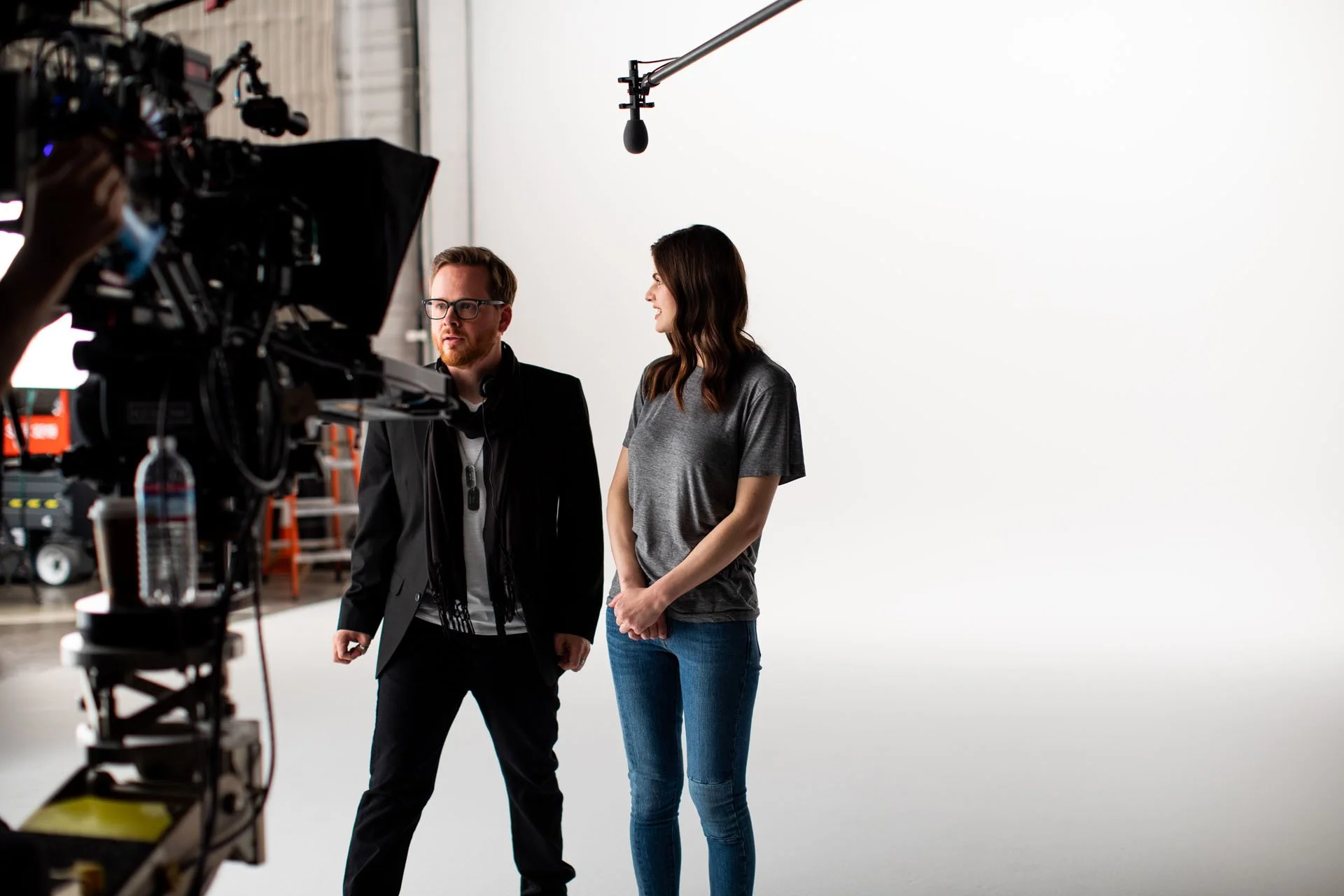 Two people, a man with glasses and a woman with long dark hair, standing on a film set in front of a white backdrop. The man is talking or explaining something, and a microphone hangs above them.