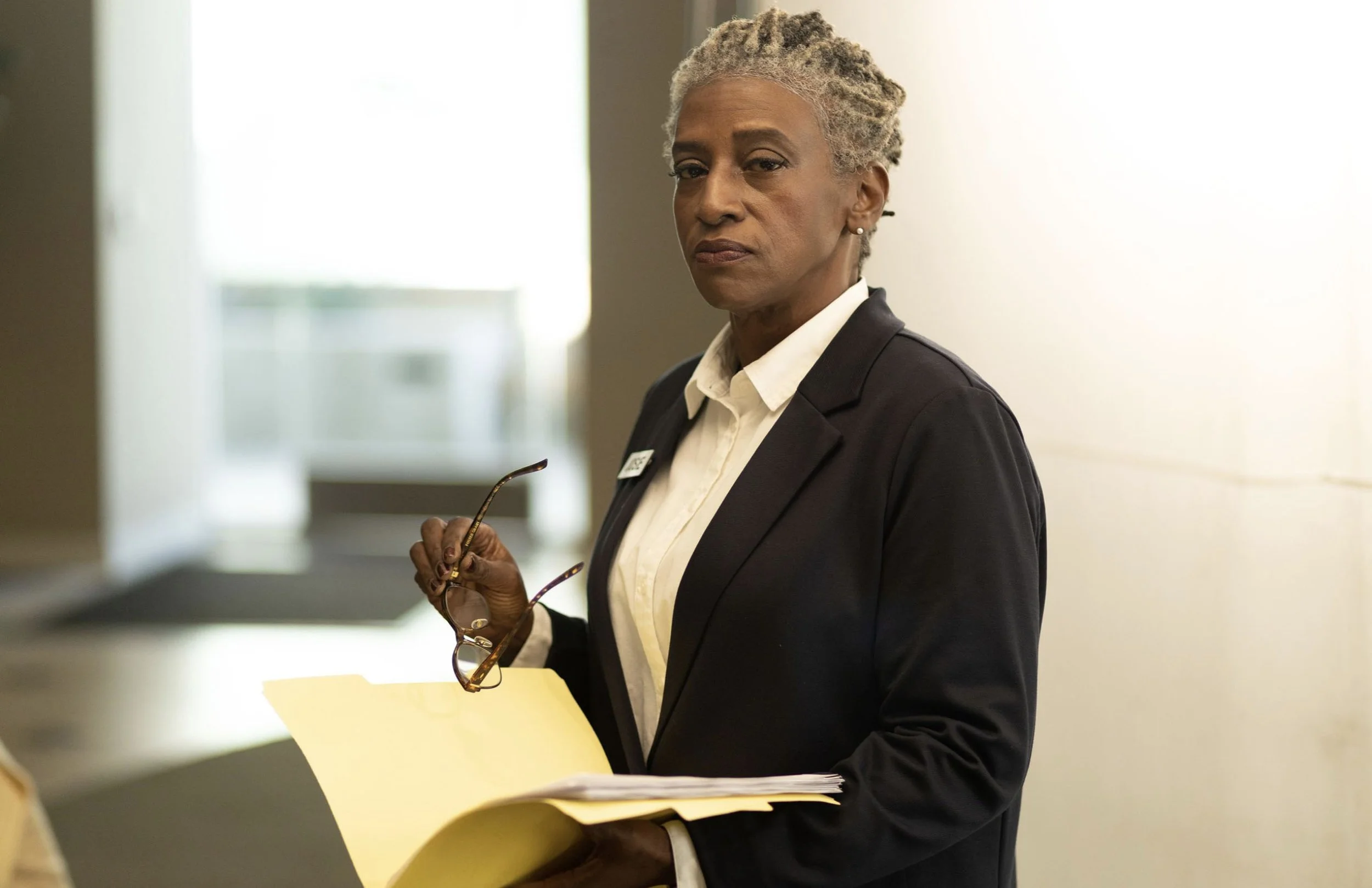 A professional woman with gray hair, wearing a black blazer and white shirt, holding a yellow folder and reading glasses, standing in a brightly lit hallway.