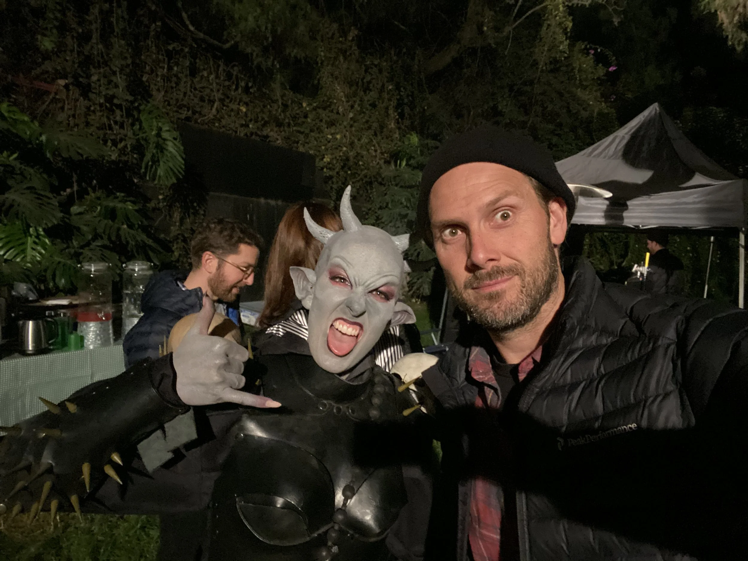 Man taking a selfie with a person in costume dressed as a fantasy creature with horns, grey skin, and spikes on arm, at an outdoor event at night.