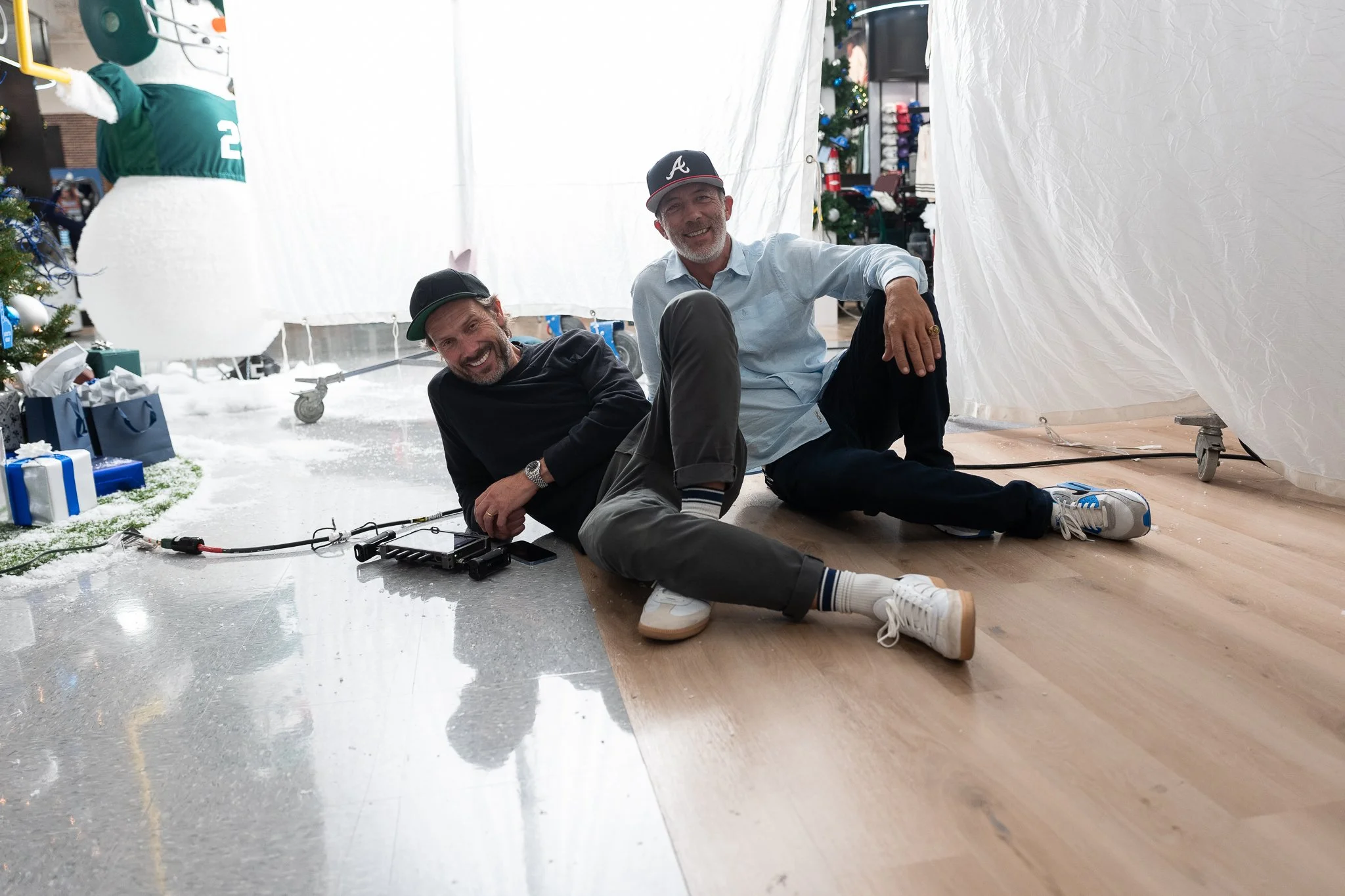 Two men sitting on the floor inside a decorated room, smiling at the camera. There is a Christmas tree with presents on the left and a white backdrop or divider behind them.