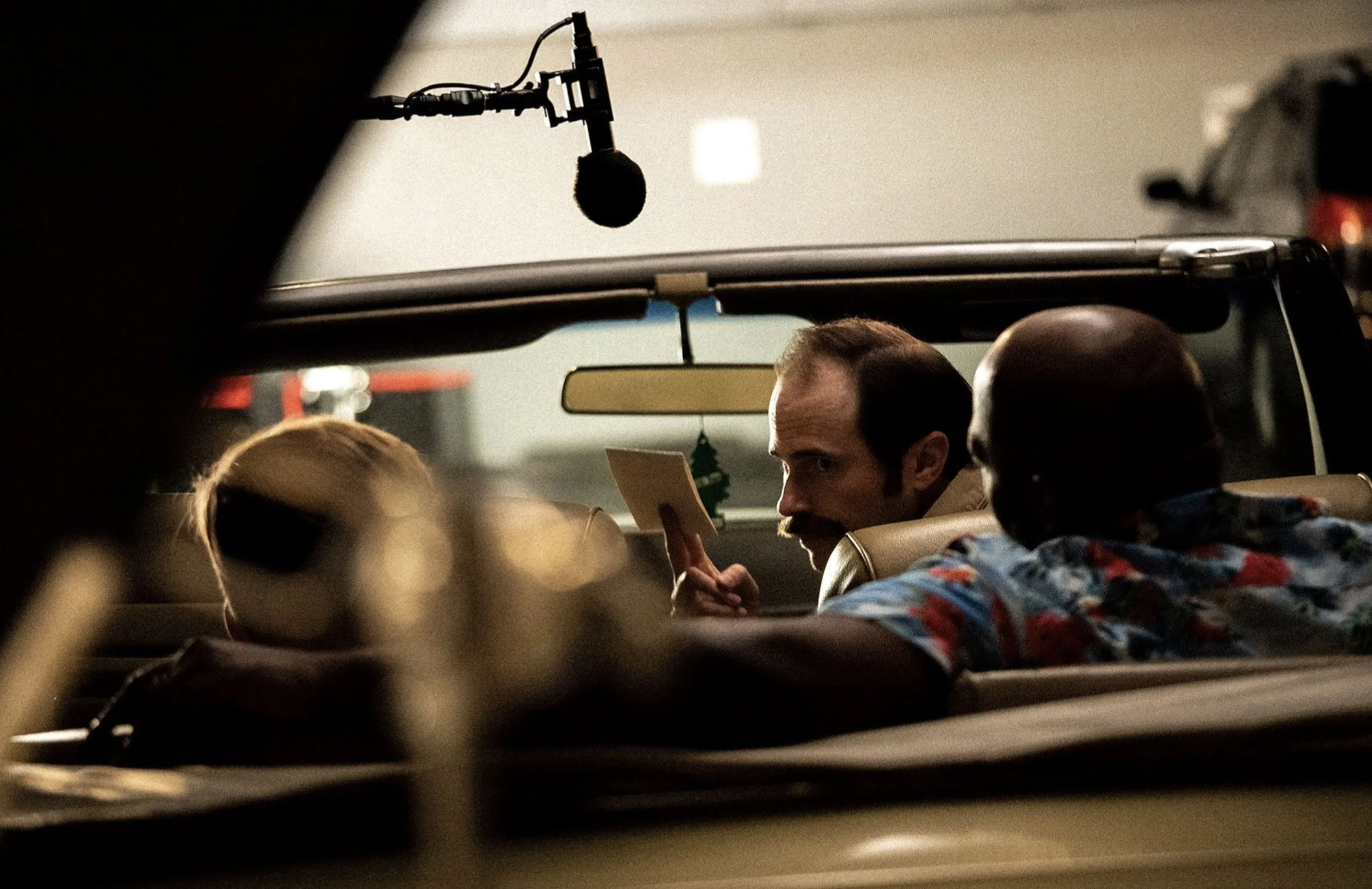 Three men sitting in a car interior, with one man holding a small notebook and looking towards the camera, while the others are partially visible and out of focus.