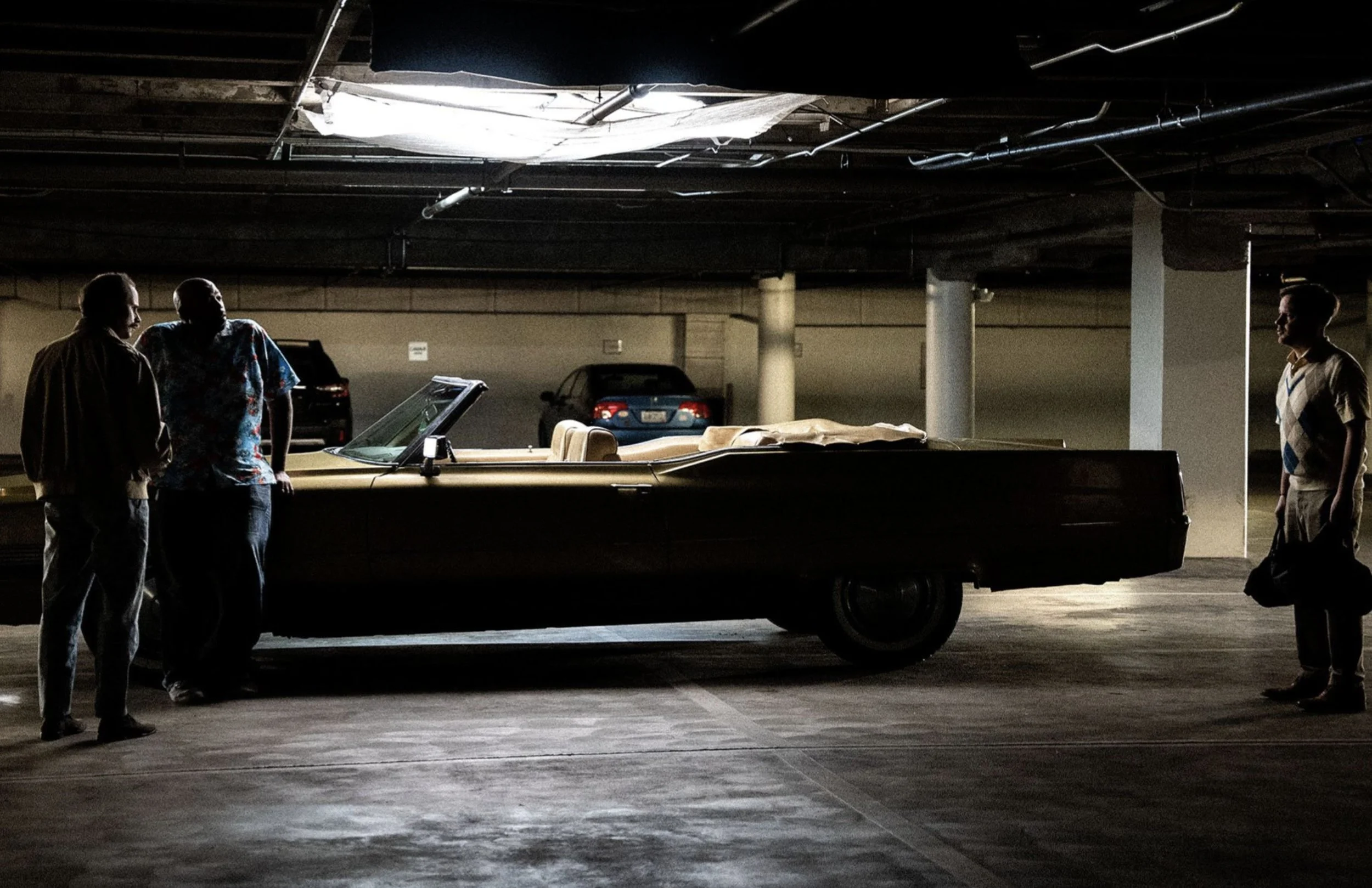 Three people standing and talking near a vintage car in a dimly lit underground parking garage with multiple parked cars in the background.
