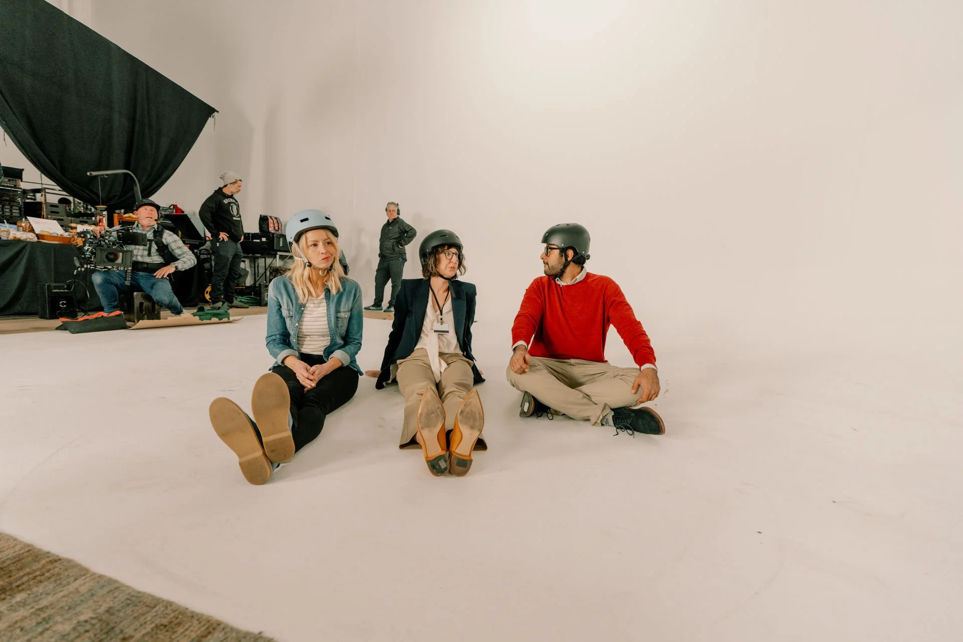 Three people sitting on the floor with helmets, having a conversation, behind them are crew members and filming equipment in a large white studio space.